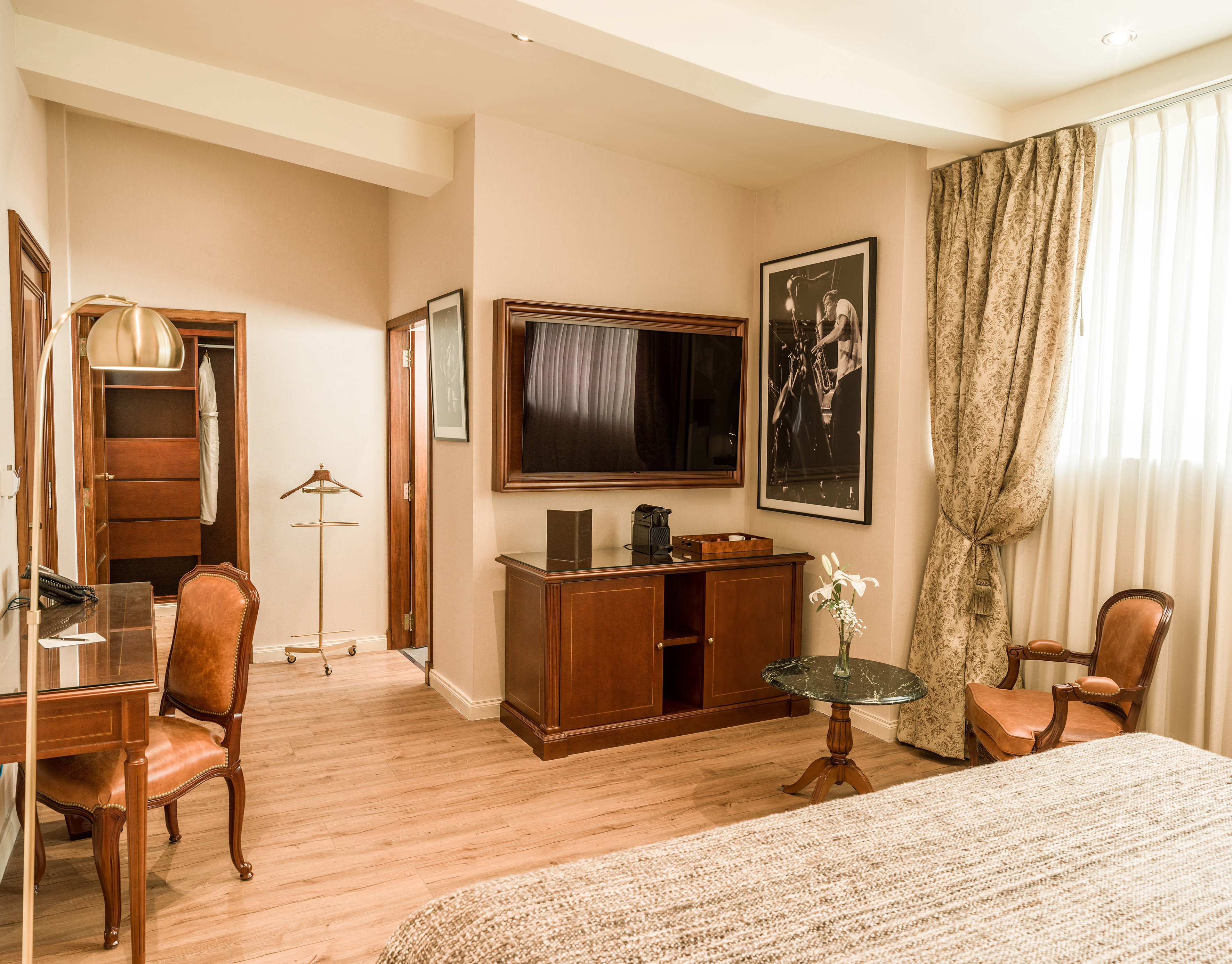 Luxurious hotel room with wood desk, TV, lounge chair, and patterned curtains.