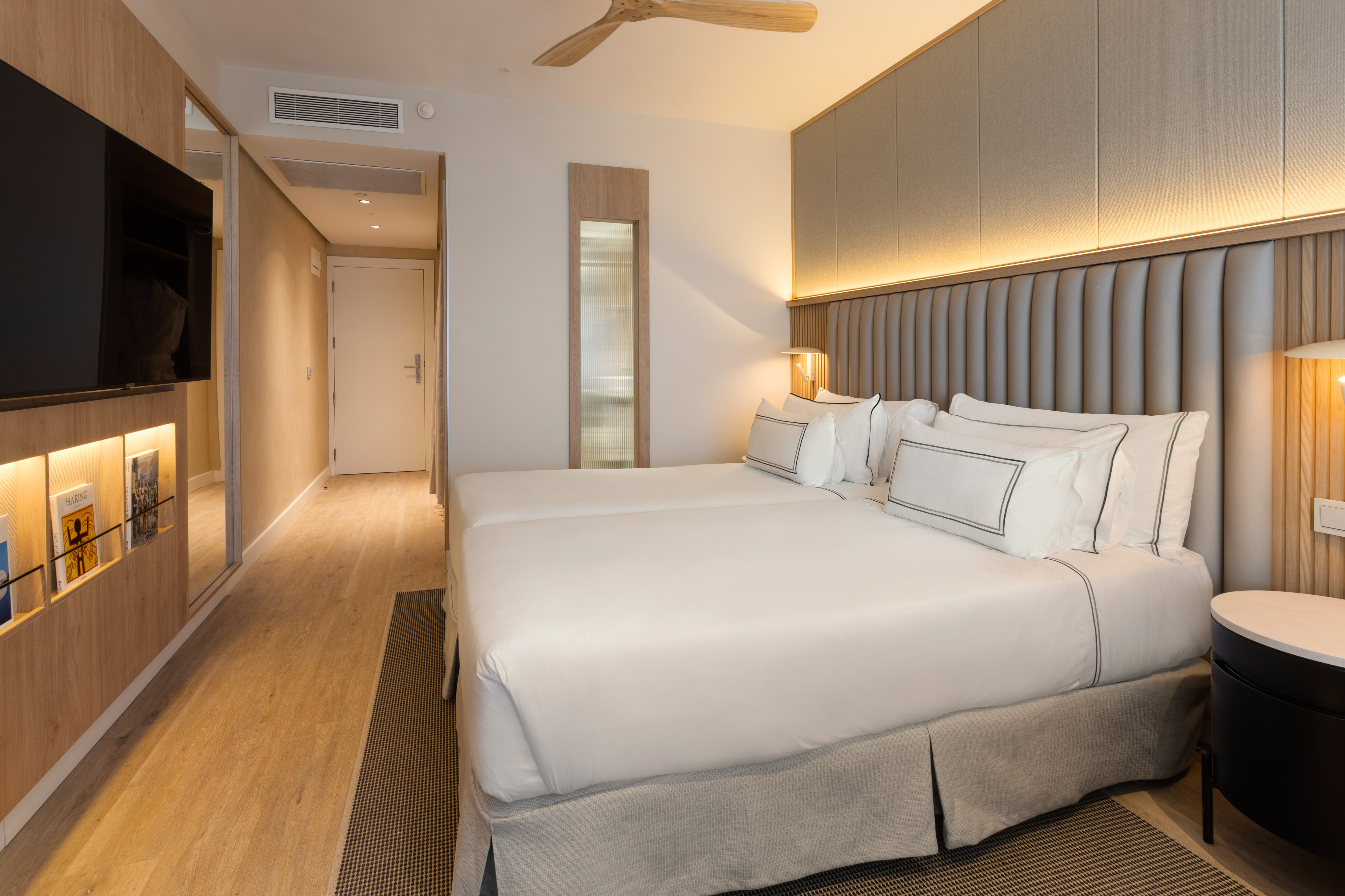 Modern hotel room with twin beds, upholstered headboard, TV, and wood details.