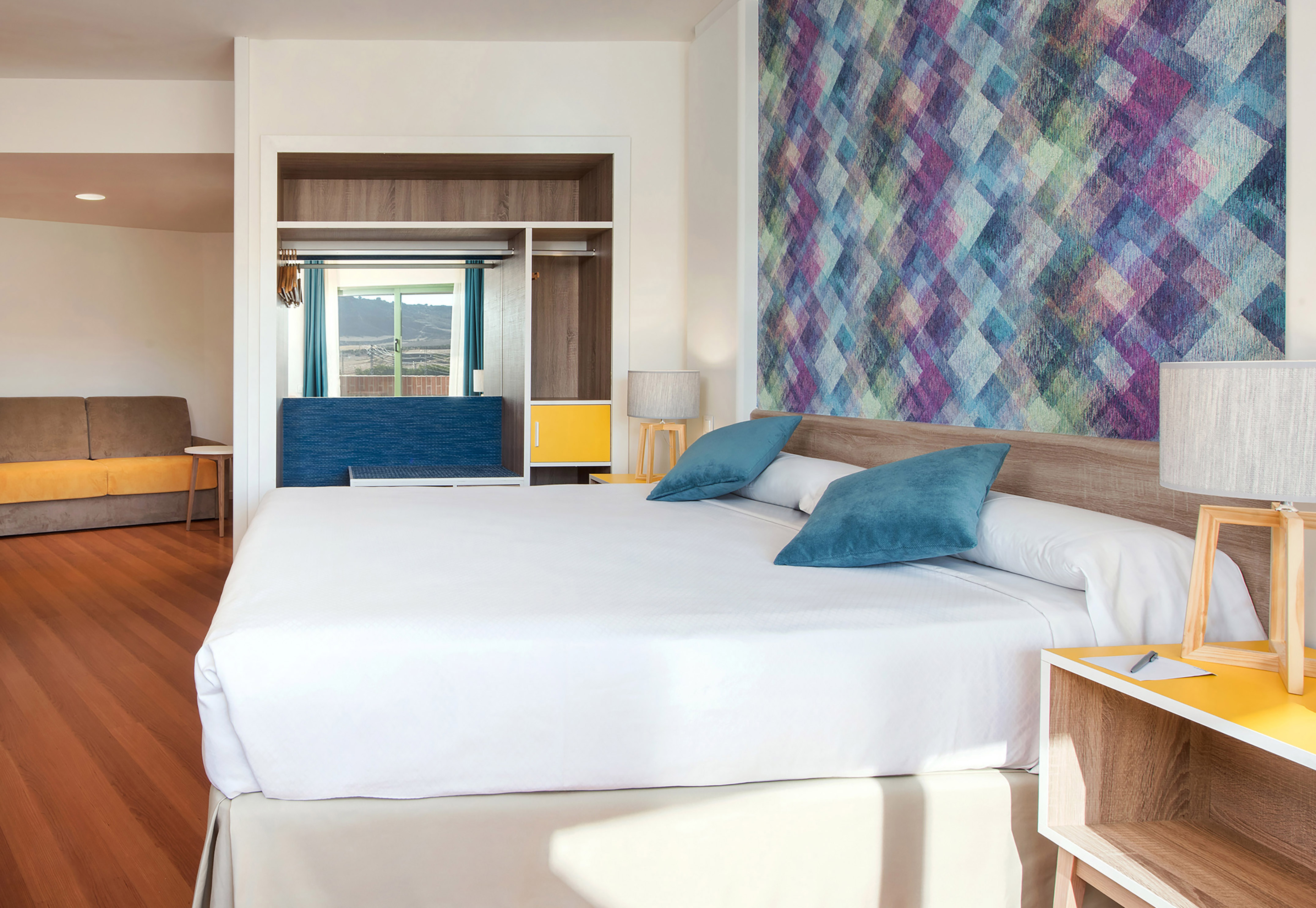 Modern hotel room with colorful geometric wall, white bed, sofa, mountain view.