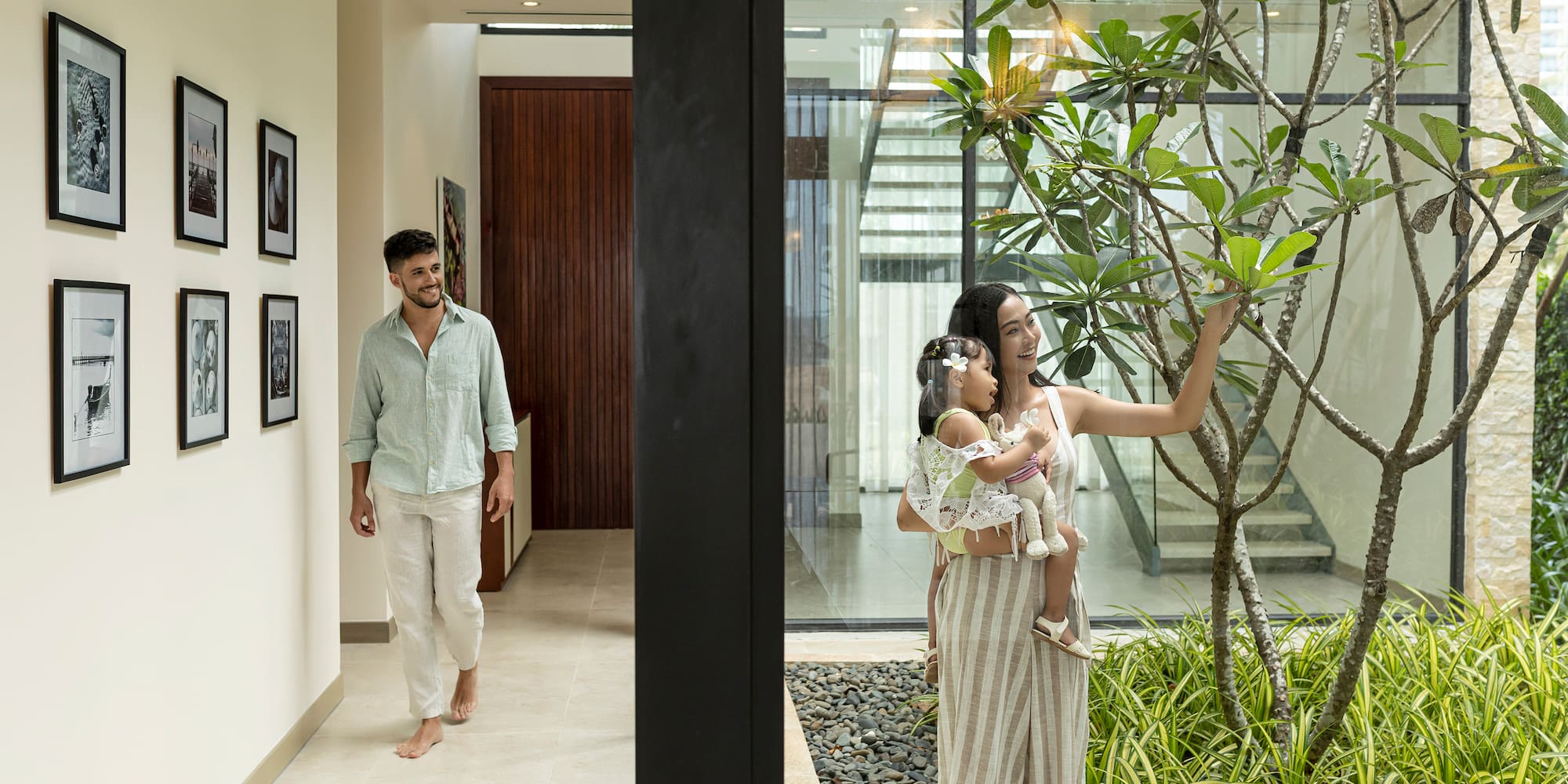a woman holding a baby and a man walking in a room with plants