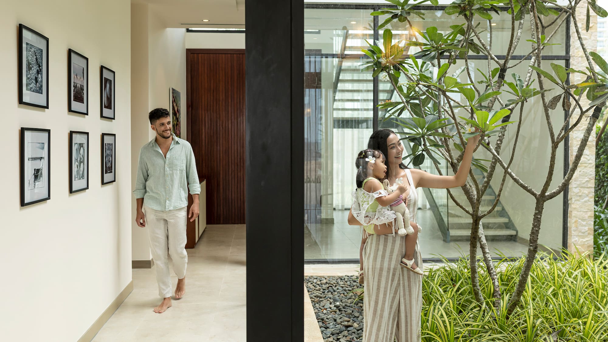 Modern home family: man in hallway, woman with baby by glass wall garden.