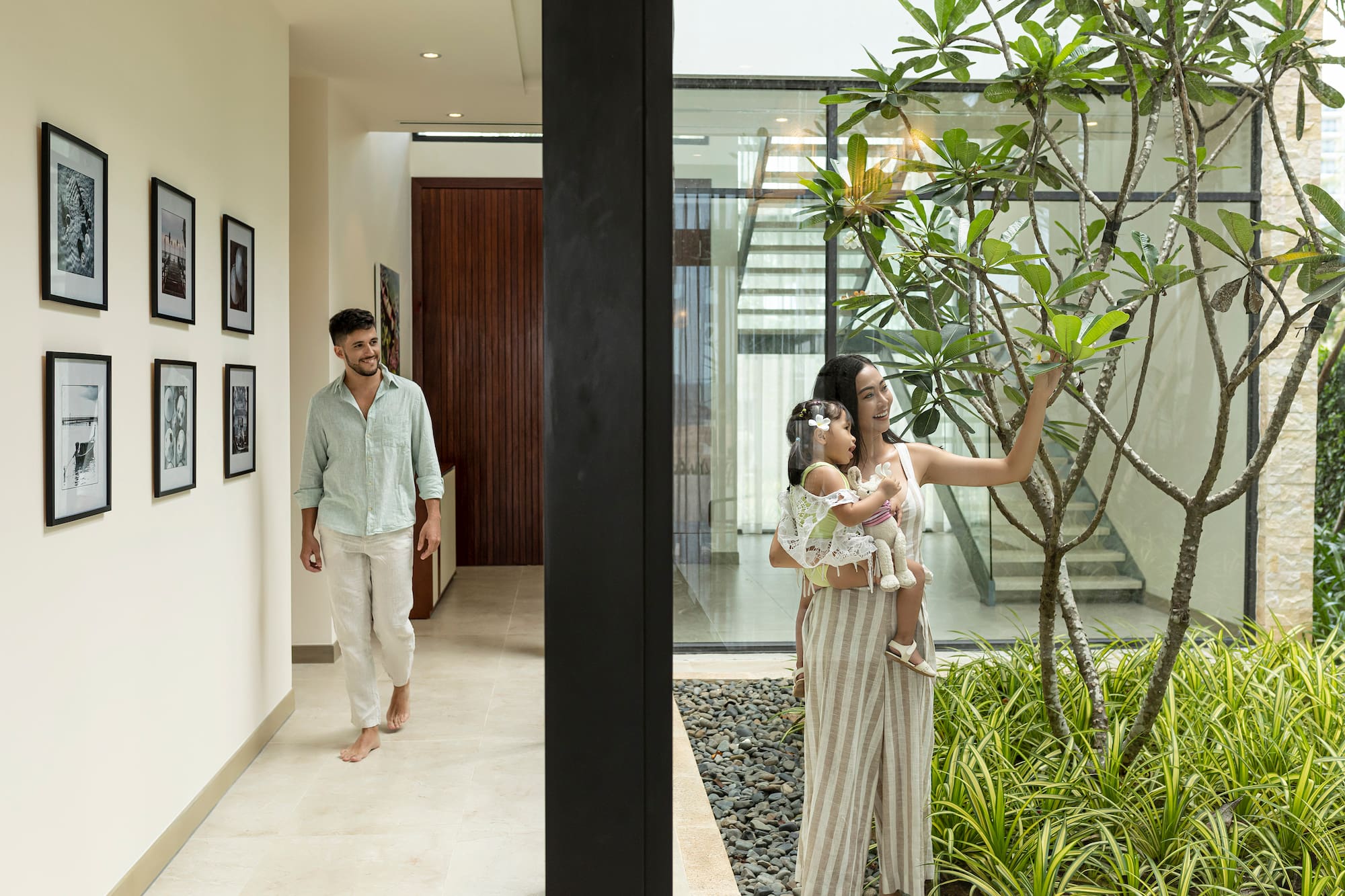 Family in modern home: man walks in hallway, woman and baby by indoor garden.