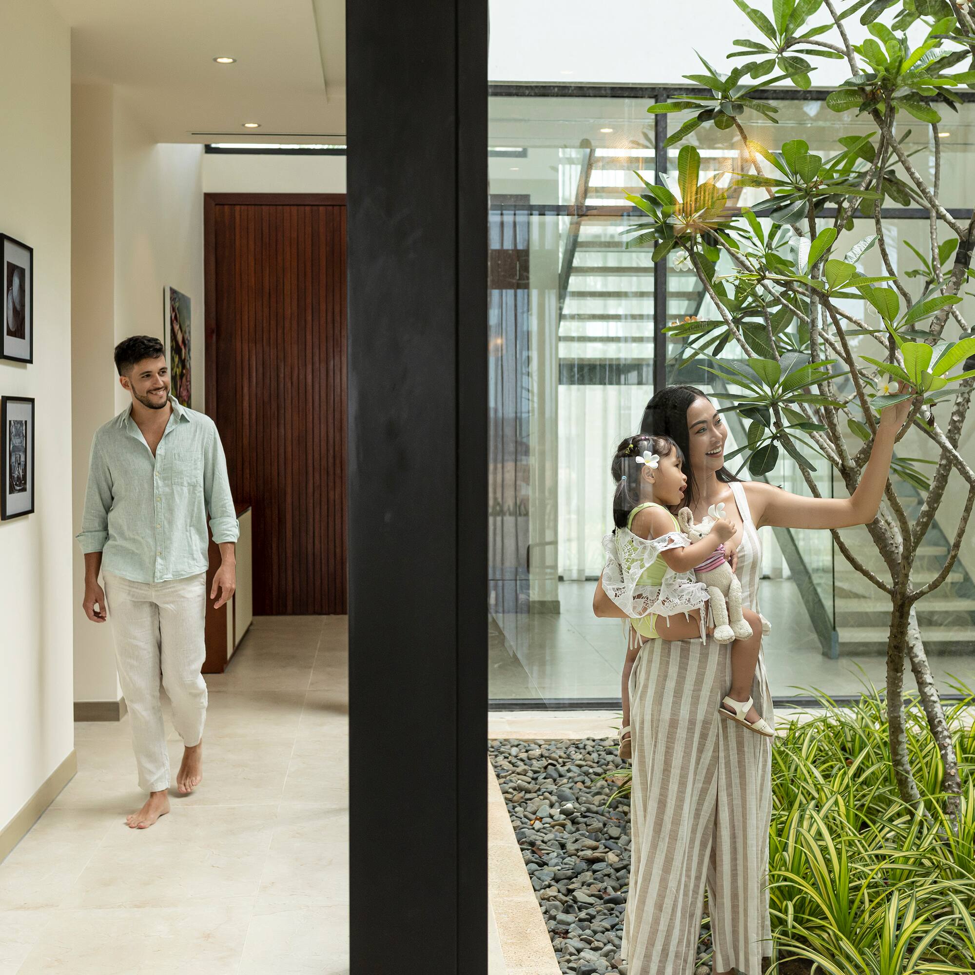 a woman holding a baby and a man walking in a room with plants
