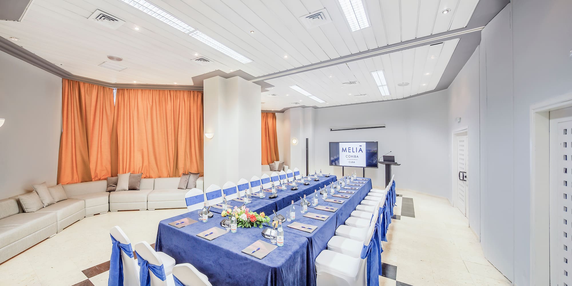 a long table with blue tablecloths and chairs in a room with a television