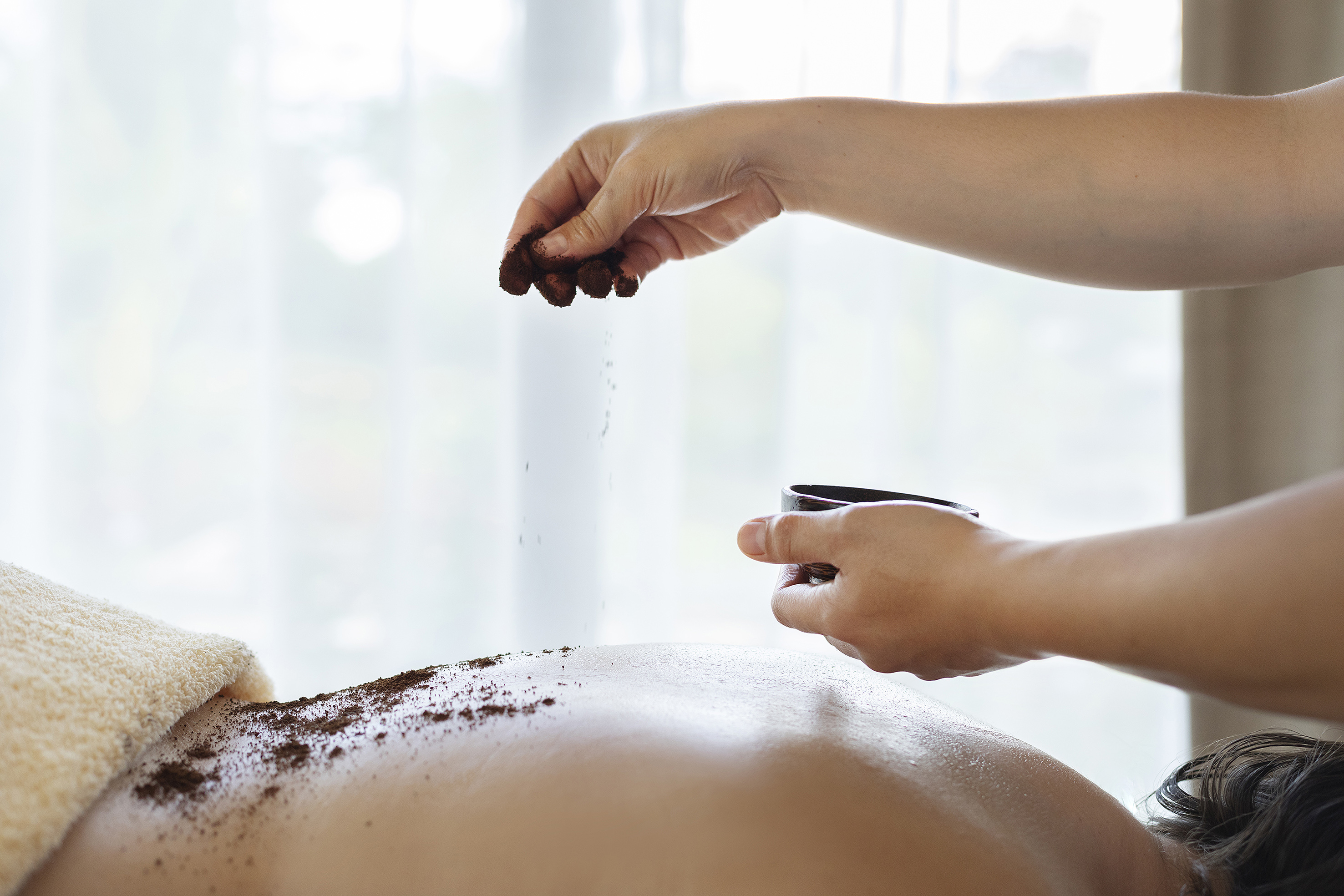 Hands applying coffee body scrub to a person's back during a spa treatment.