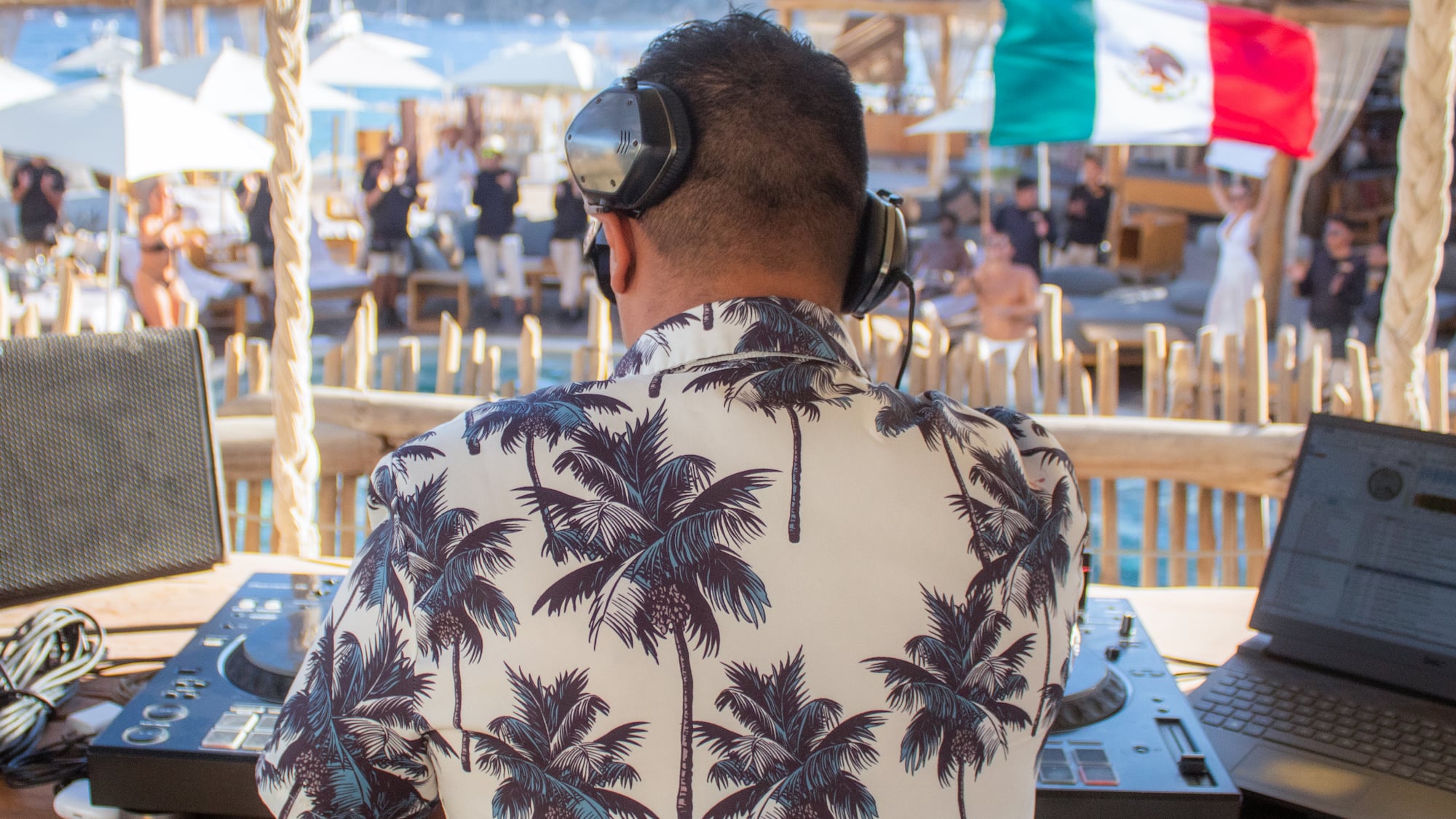 DJ in palm shirt with headphones at a lively Mexican beach club party.