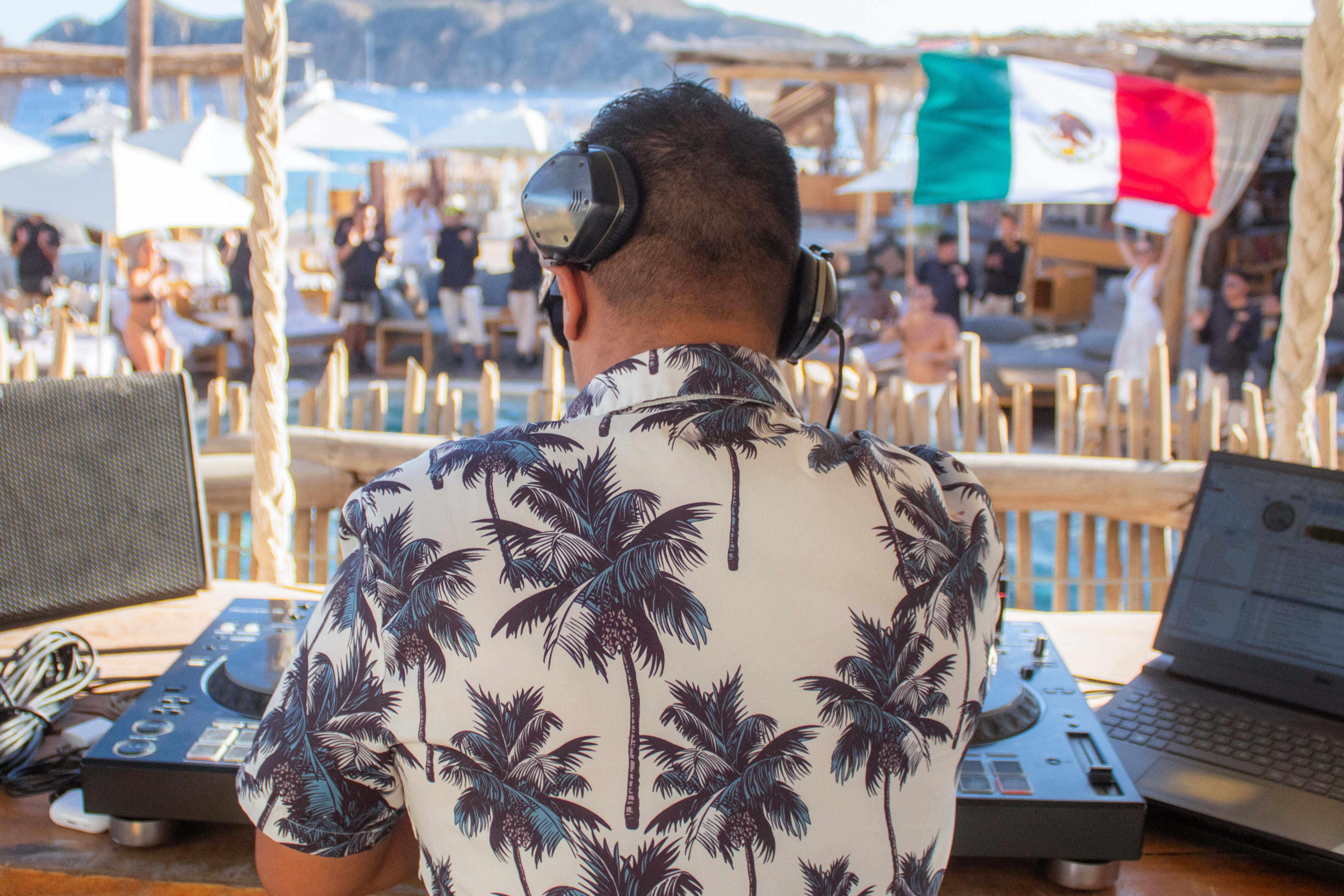 DJ in palm shirt with headphones at a lively Mexican beach club party.