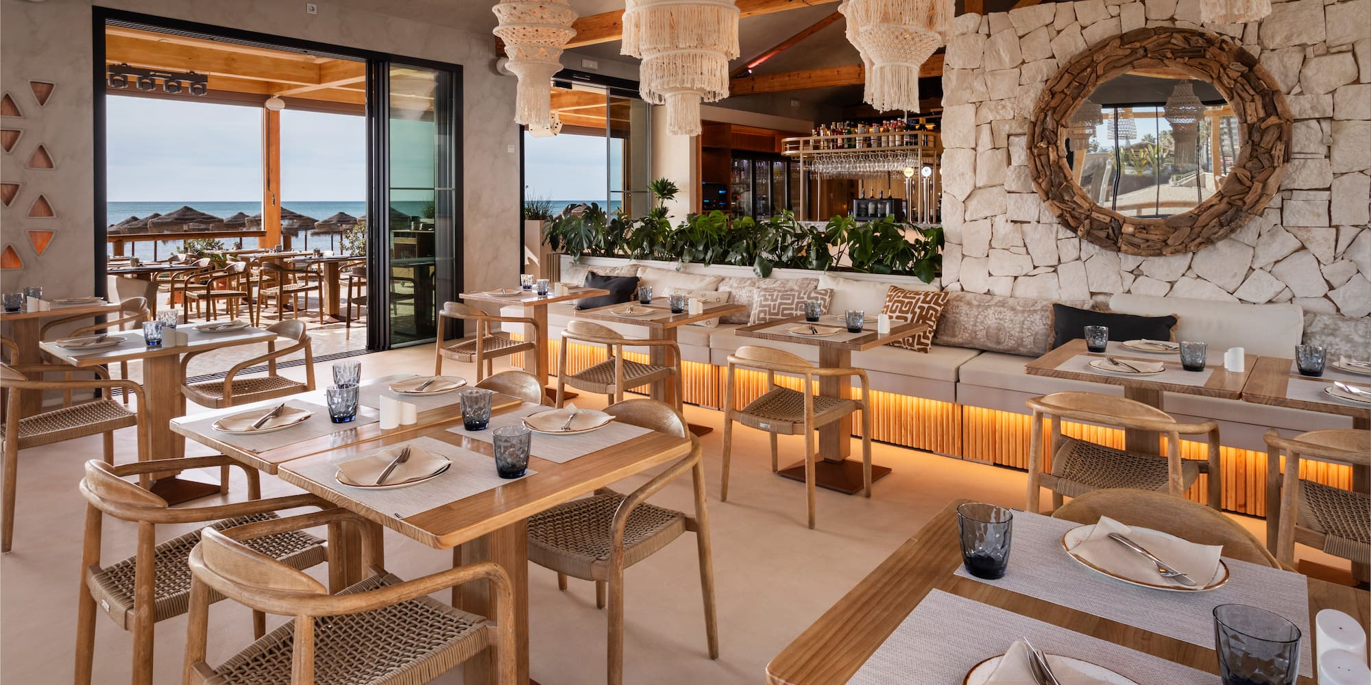 Beachfront restaurant with macrame chandeliers, wooden tables, and sea view.