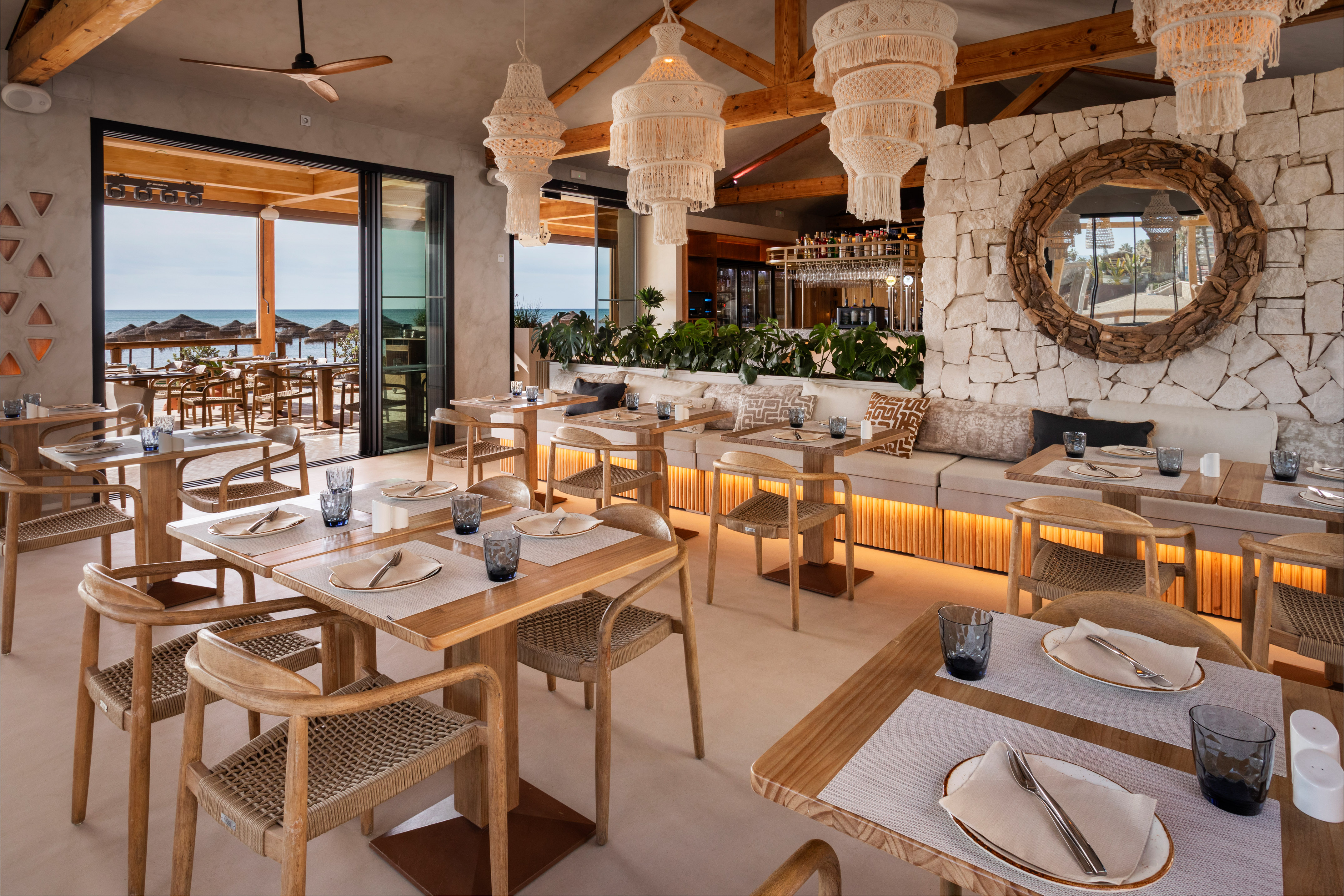 Beachfront restaurant with macrame chandeliers, wooden tables, and sea view.
