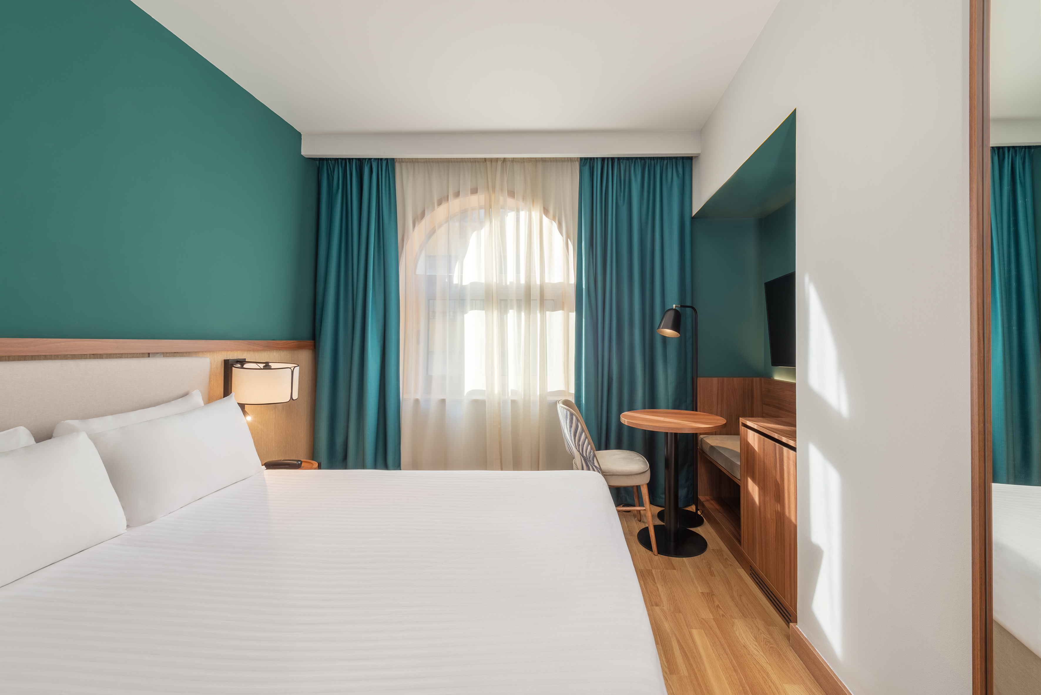 Clean hotel room with white bed, teal wall, arched window, and wood floor.
