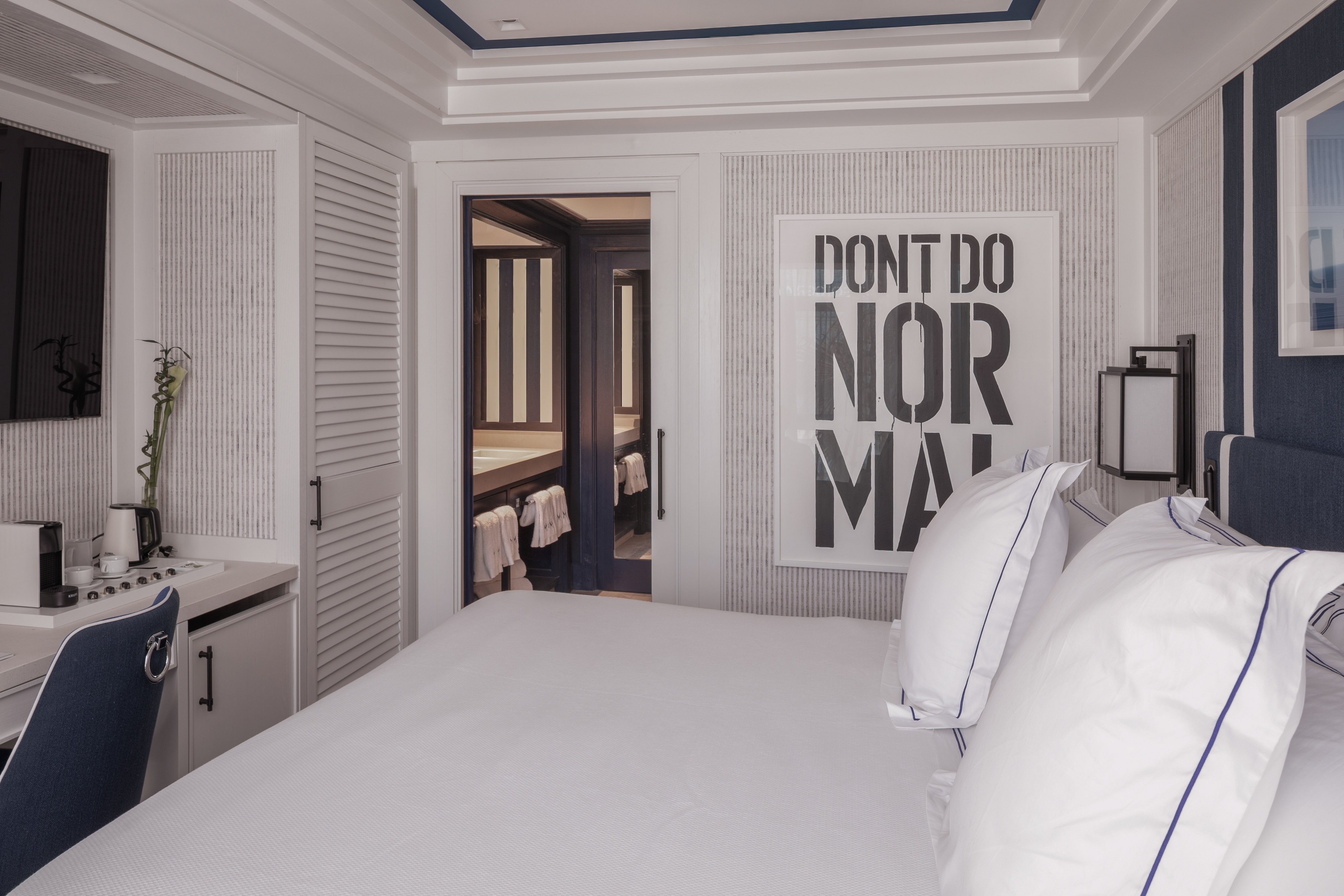 Modern hotel bedroom with white bed, blue details, and 'DONT DO NORMAL' art.