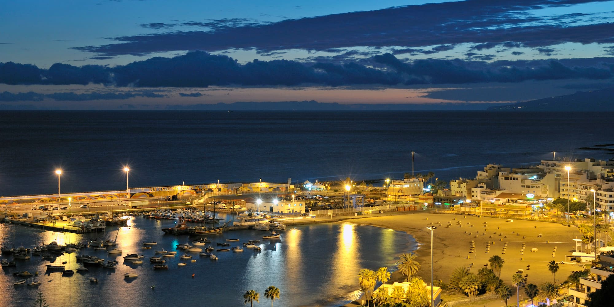 Vista nocturna de puerto, playa, palmeras y ciudad costera iluminada.
