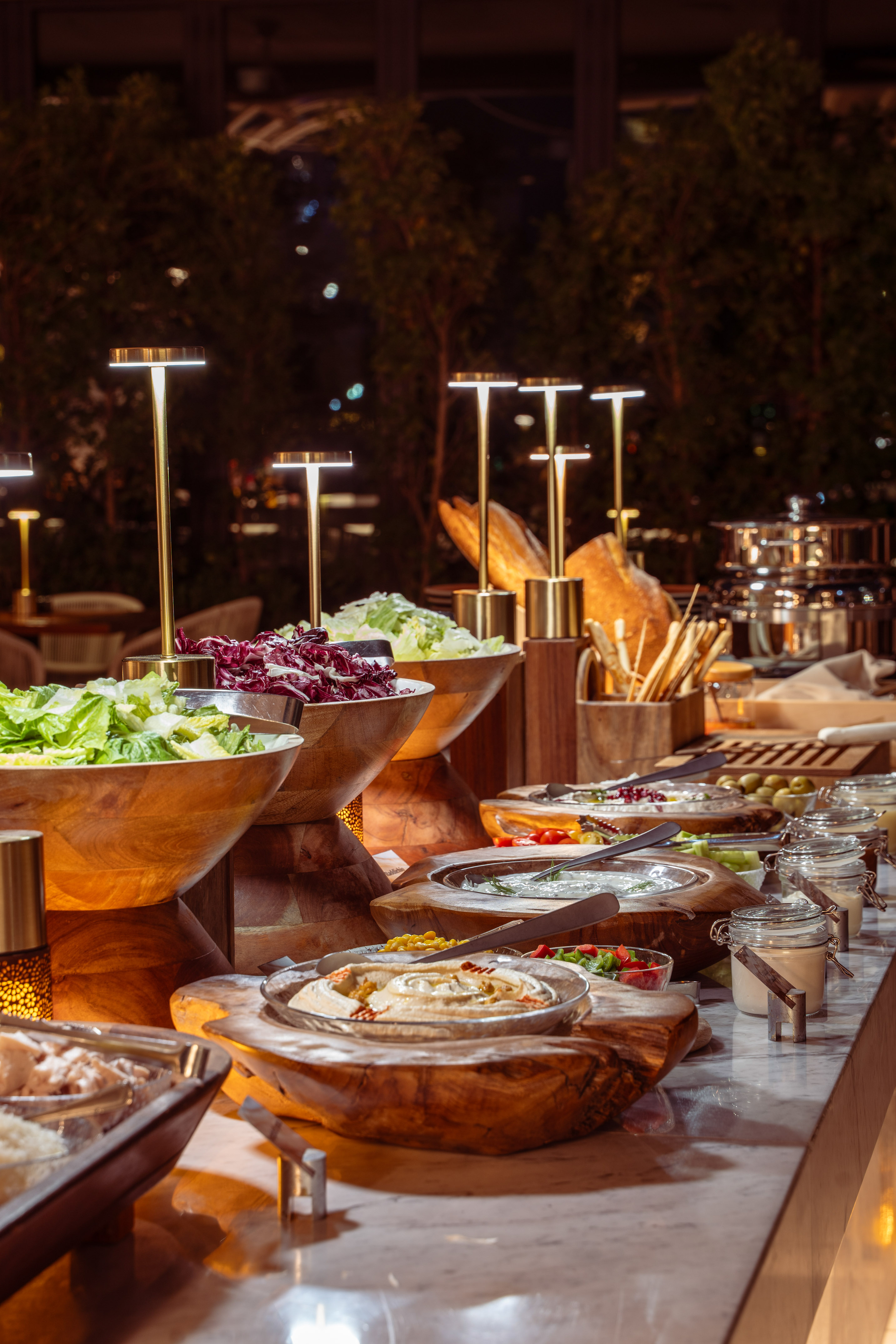 Elegant buffet with fresh salads, hummus, dips, and bread under warm lights.