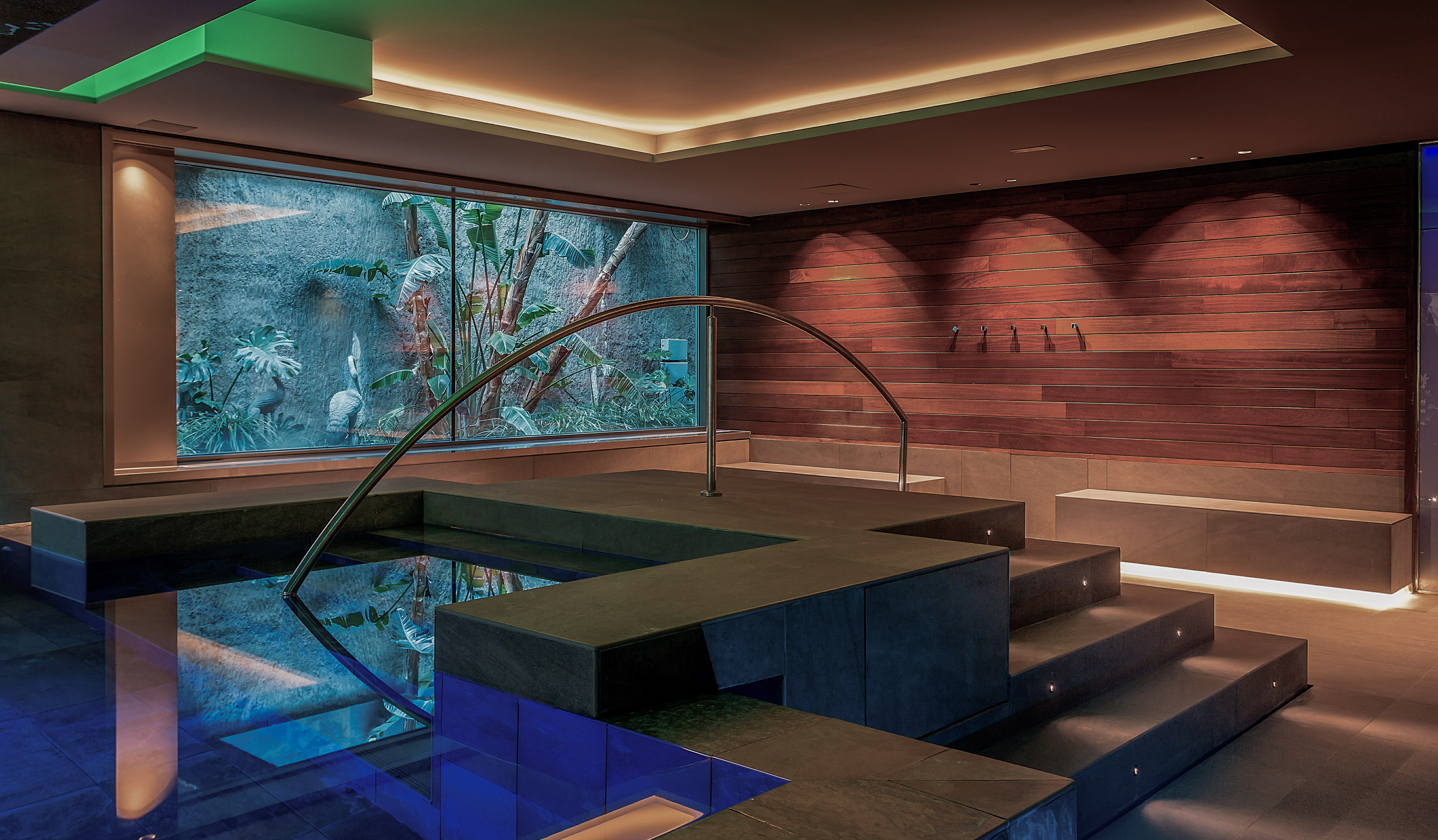 Modern indoor spa pool with blue light, steps, and tropical window view.
