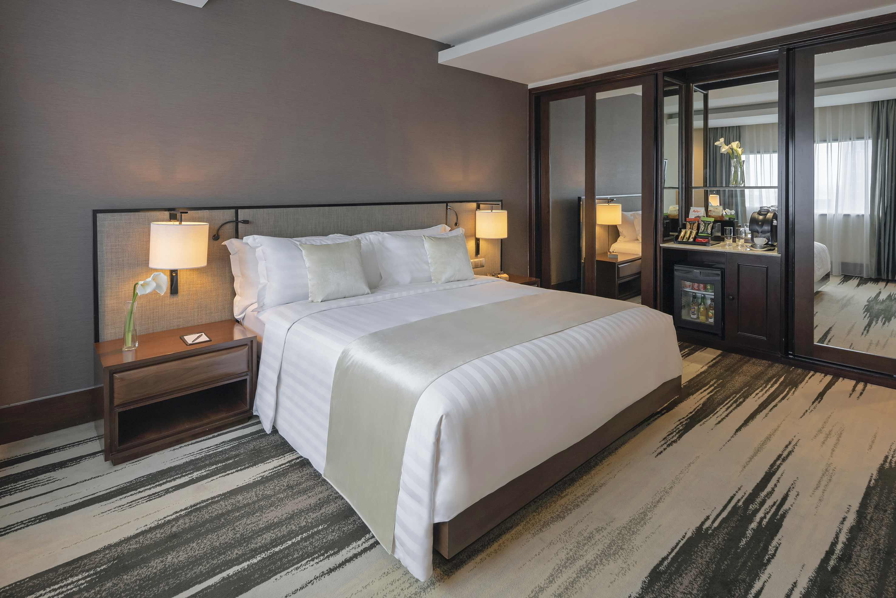 Modern hotel bedroom with king bed, nightstands, and mini-bar.