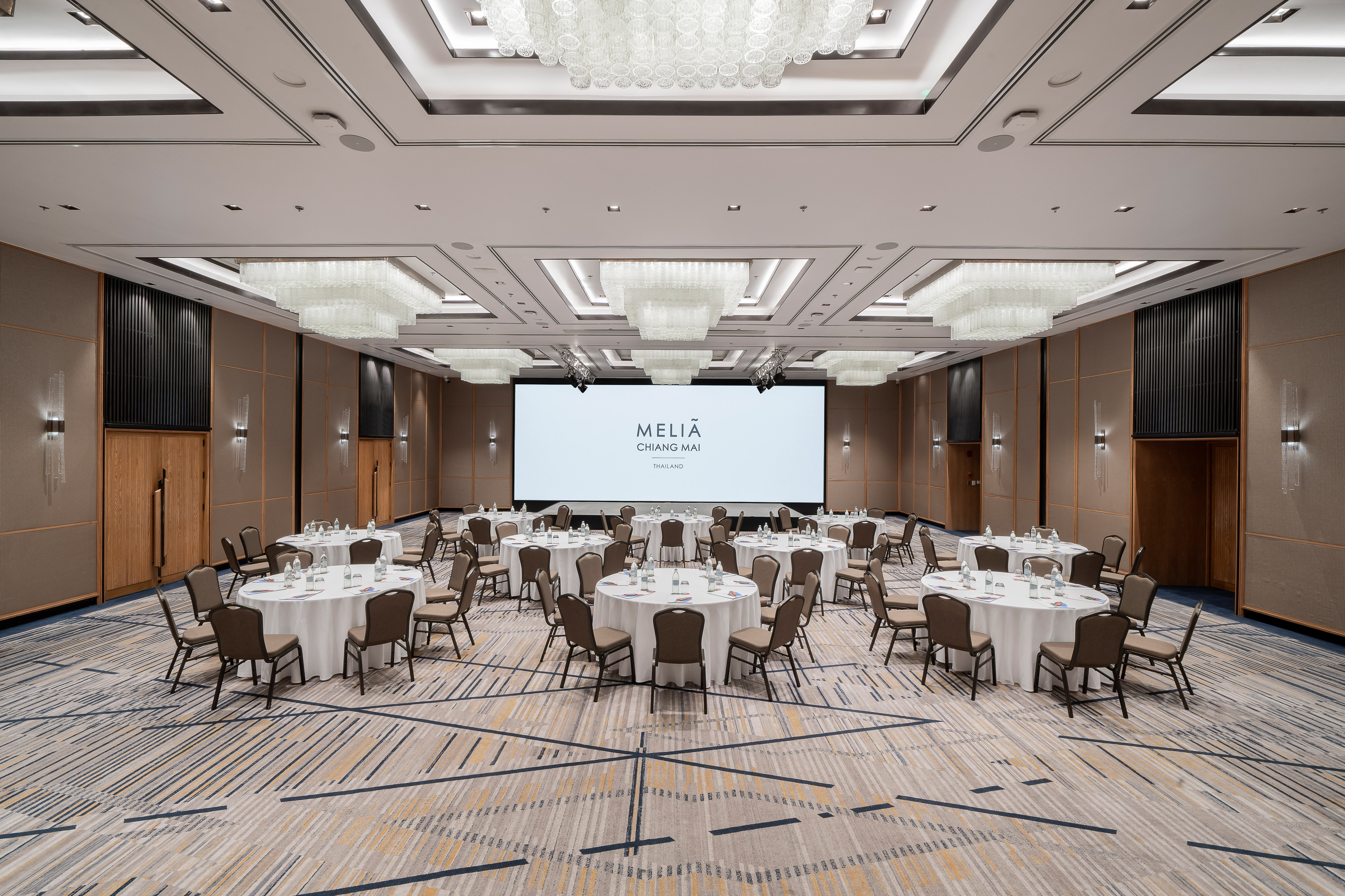 Meliã Chiang Mai ballroom with round tables, large screen, and chandeliers.