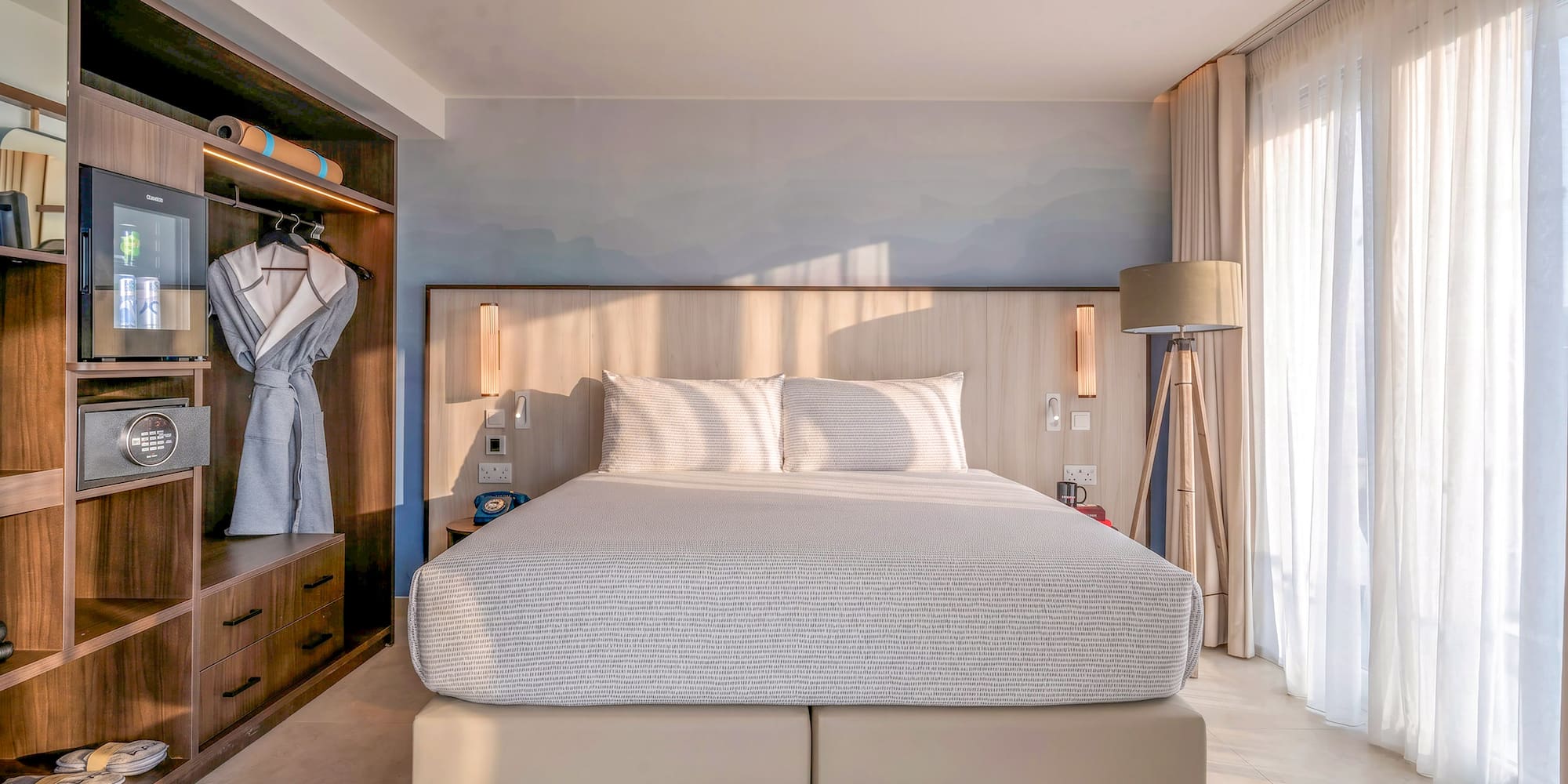 Bright hotel room with king bed, built-in storage, and sheer curtains.