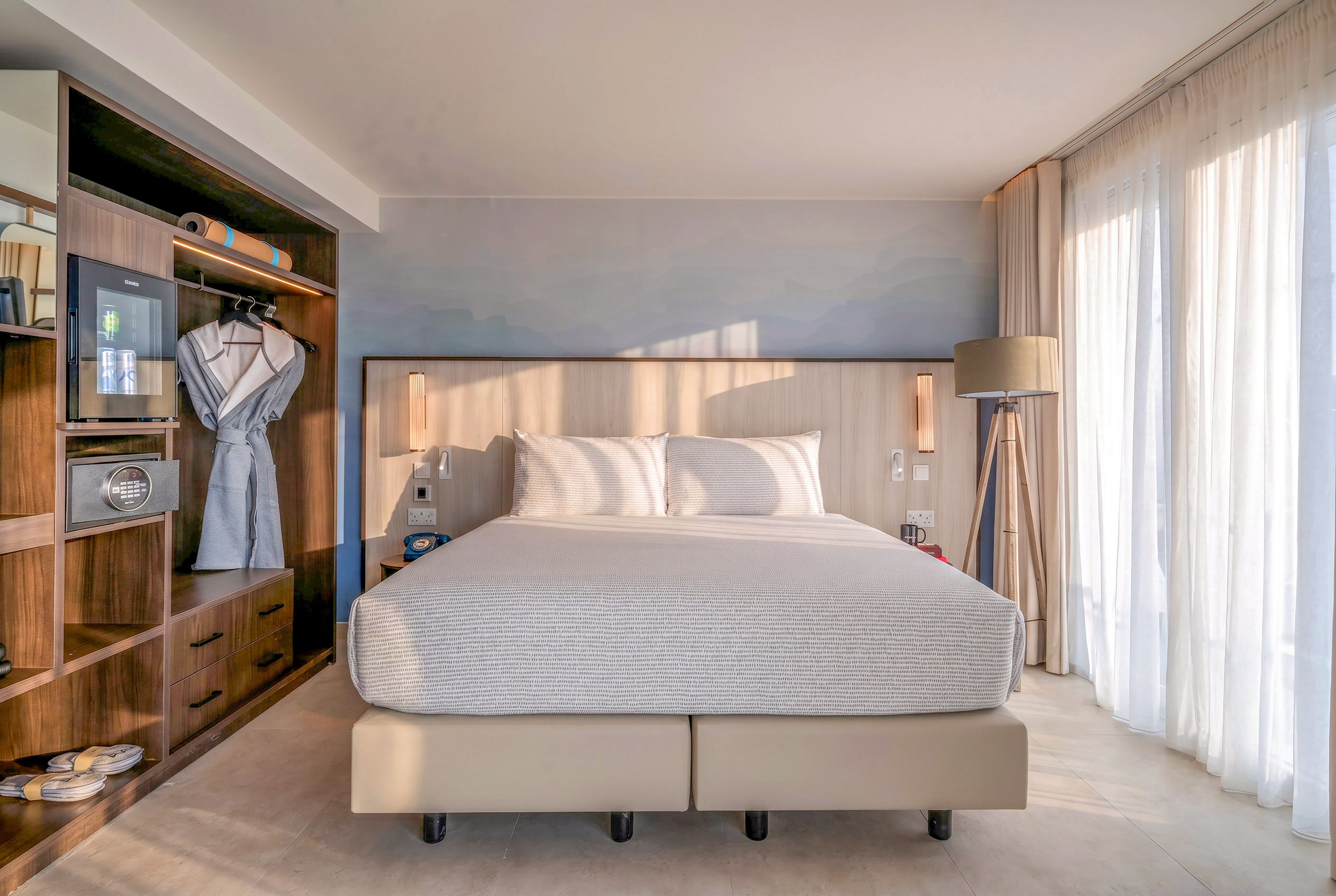 Bright hotel room with king bed, built-in storage, and sheer curtains.
