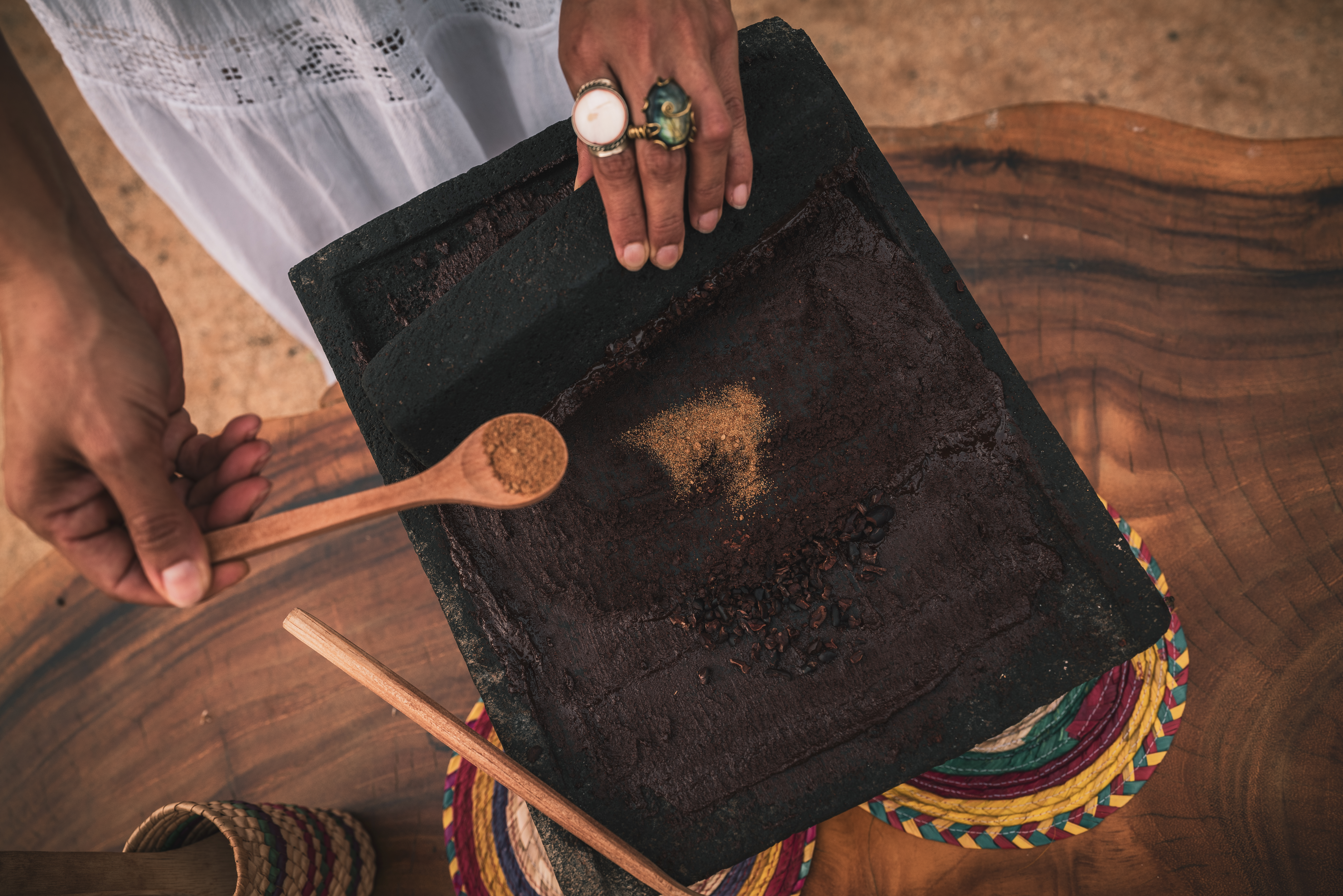 Hands grinding cacao beans and spices on a metate with a wooden spoon.