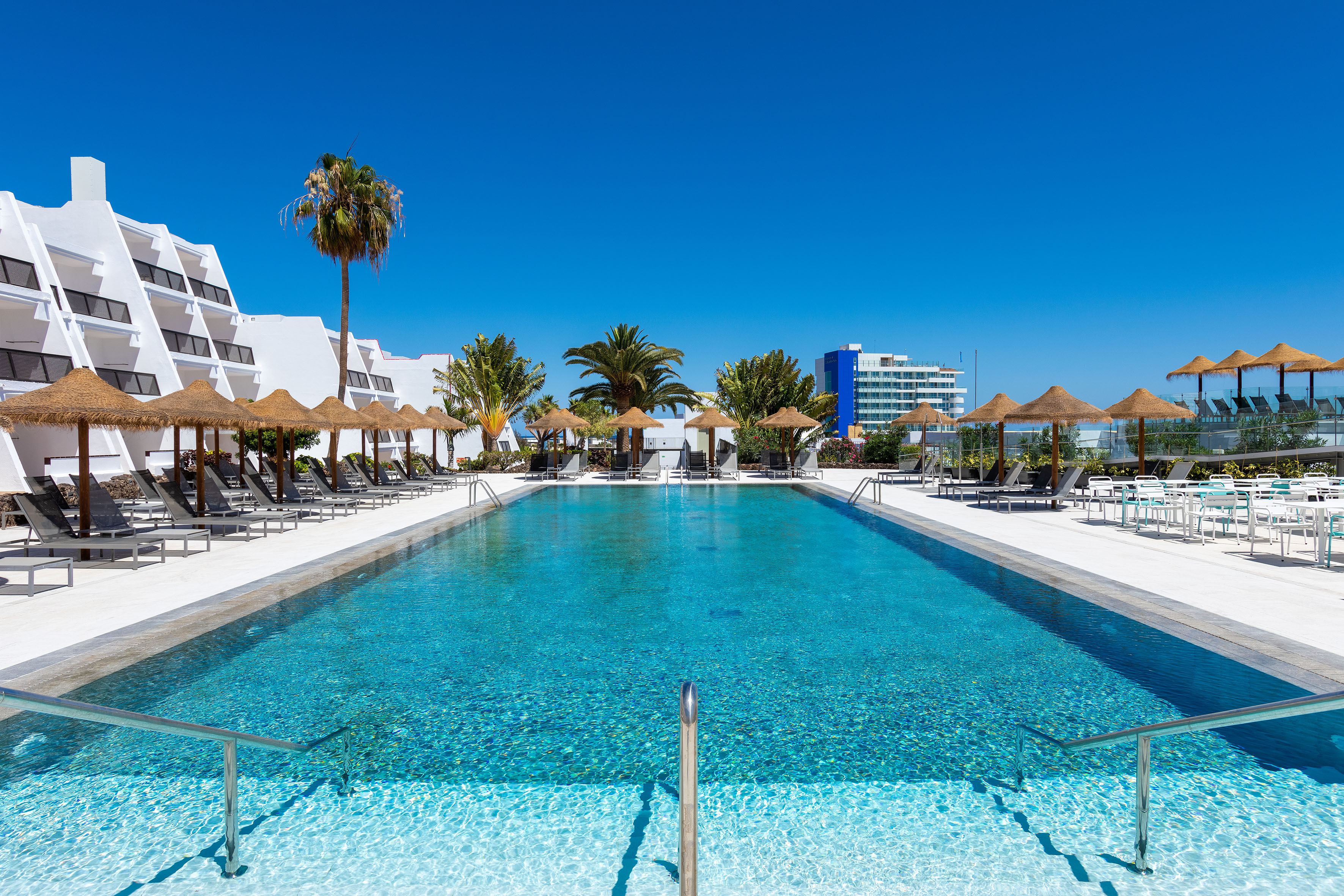 Sunny resort pool with palm trees, straw umbrellas, and hotel building.