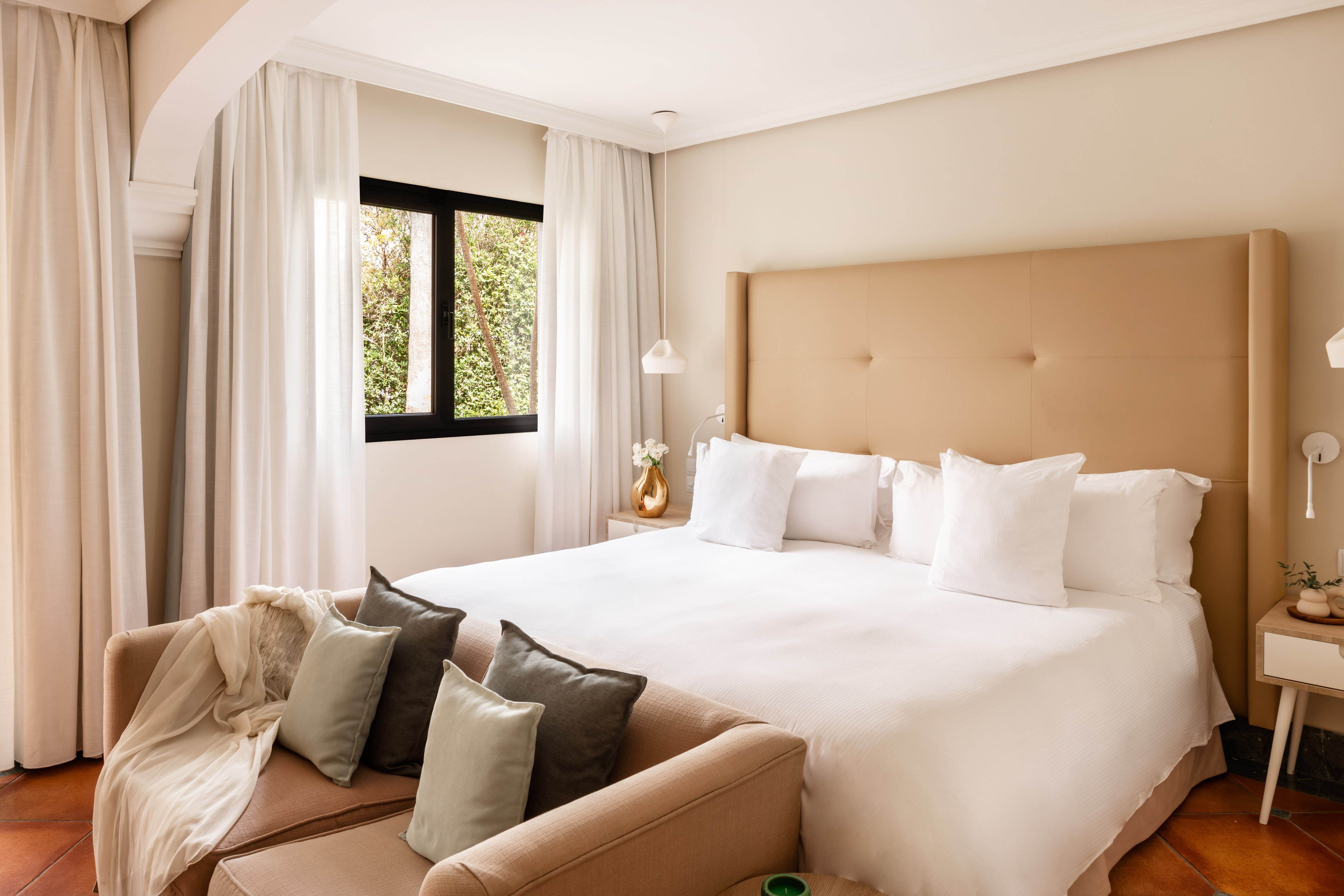 Bright bedroom with beige headboard, white bed, cozy sofa, and window.