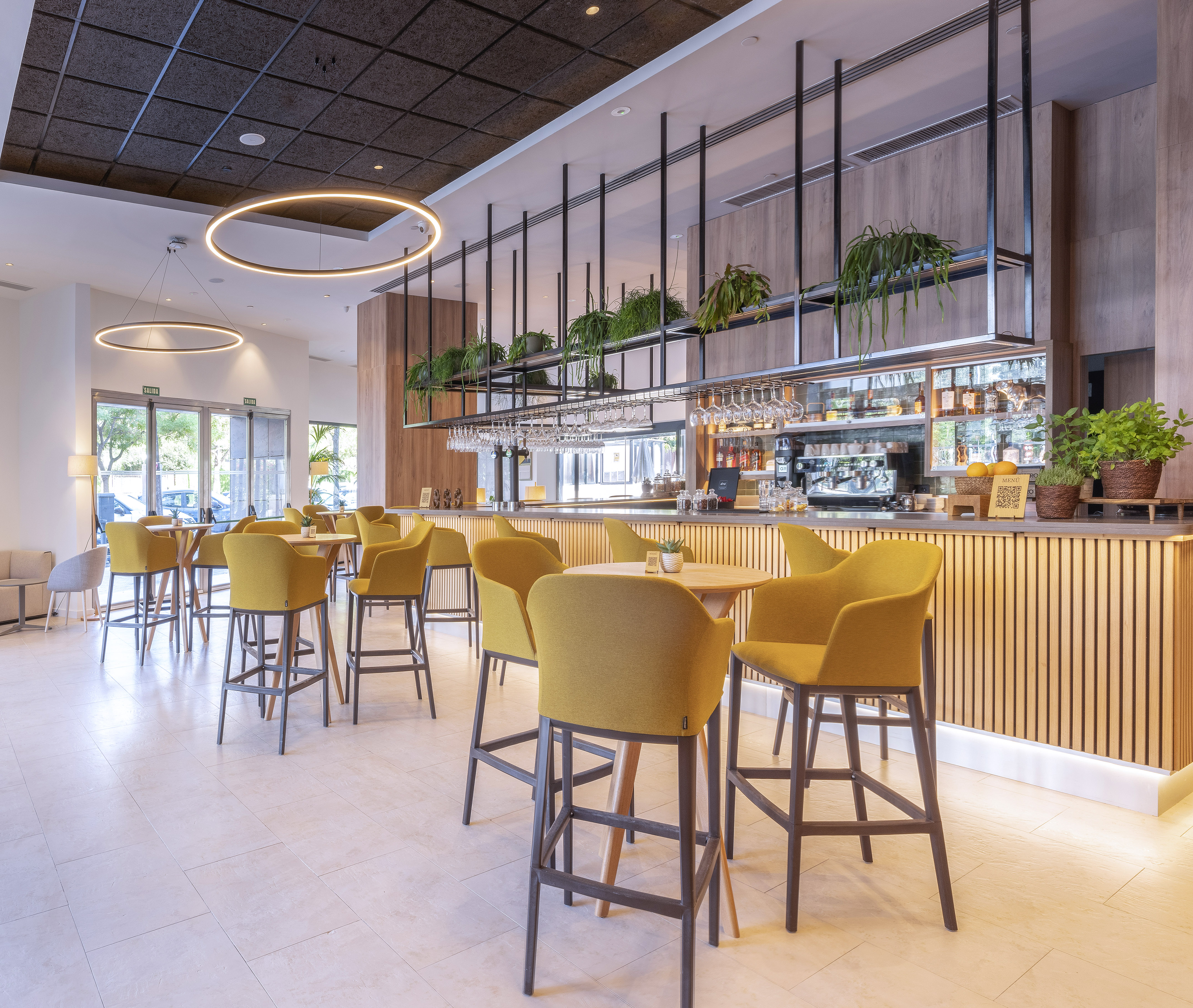 Modern hotel bar with yellow upholstered bar stools, wooden counter, and greenery.