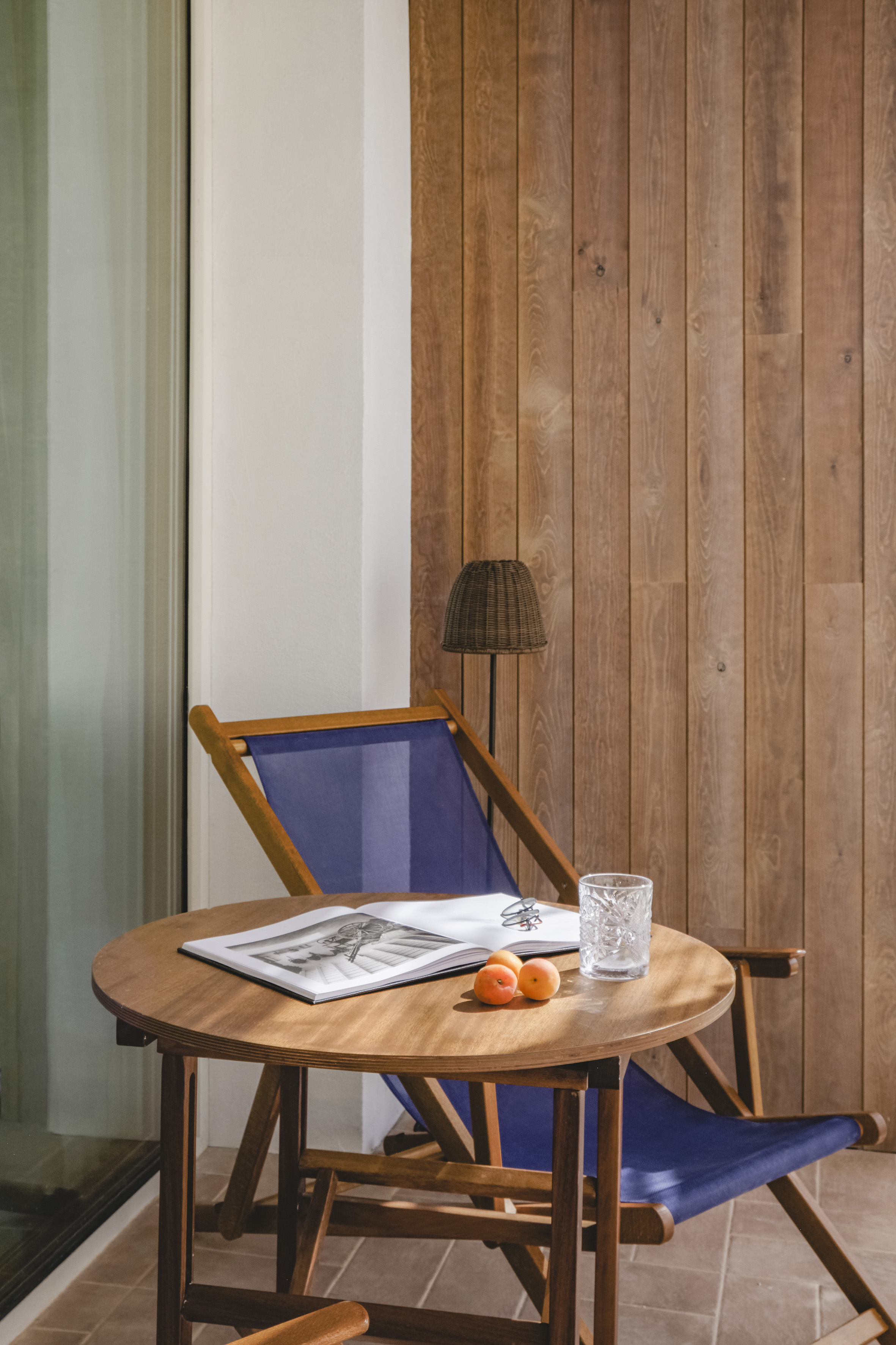 Cozy balcony: wooden table with book, apricots, blue director chair, wood wall.