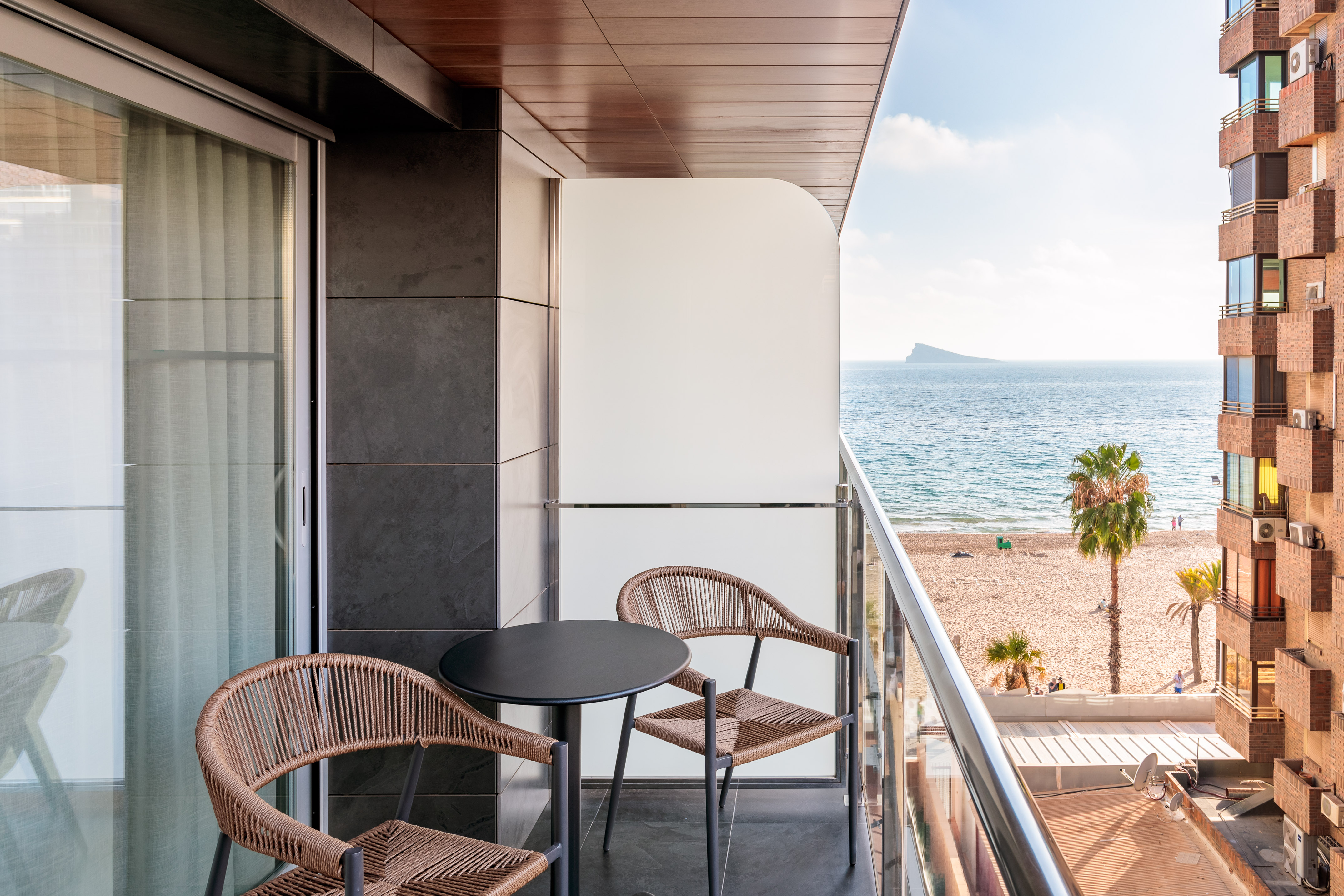 Balcony with patio chairs, ocean view, sandy beach, palm trees, distant island.