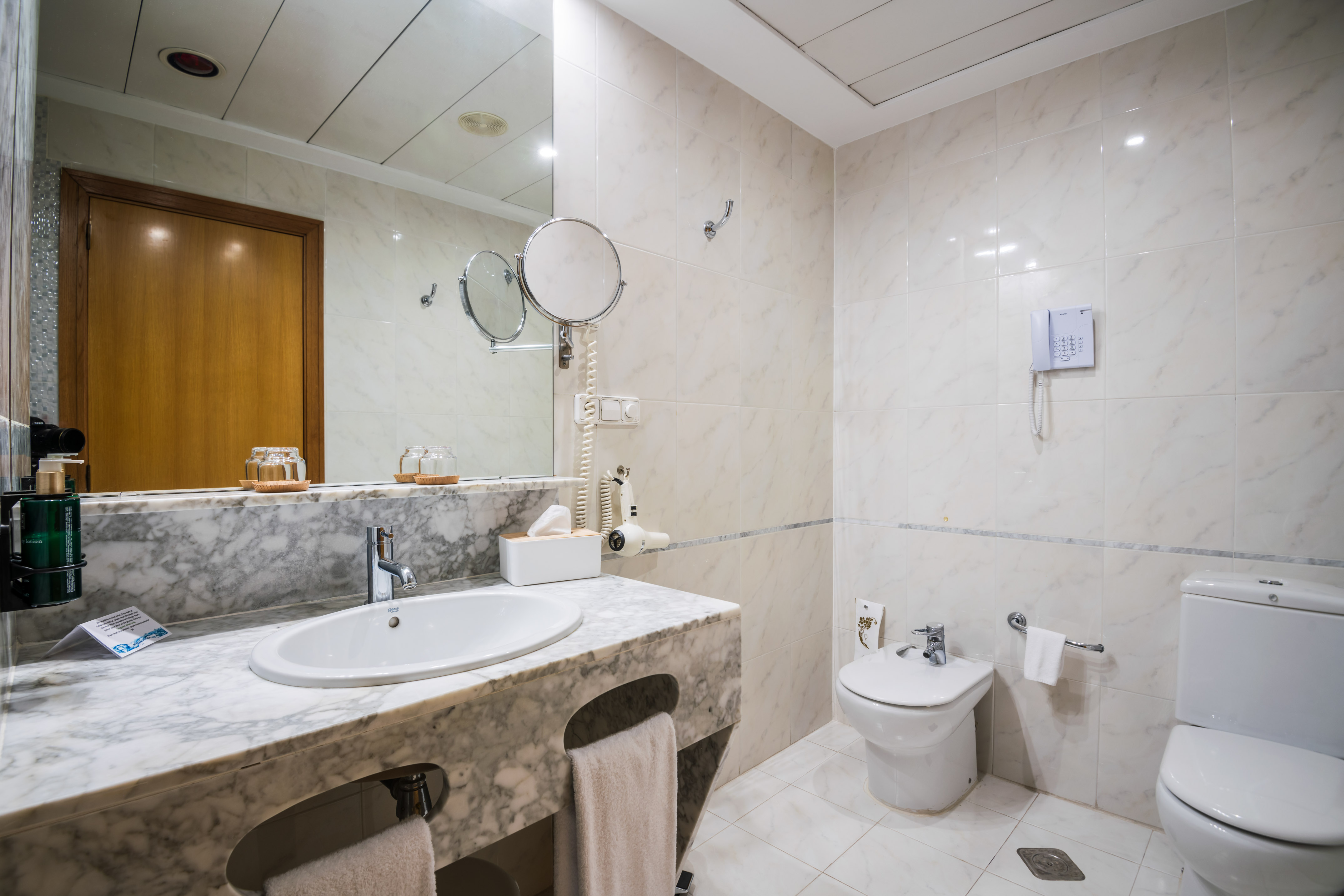 White and marble hotel bathroom with vanity, sink, toilet, bidet, and mirror.