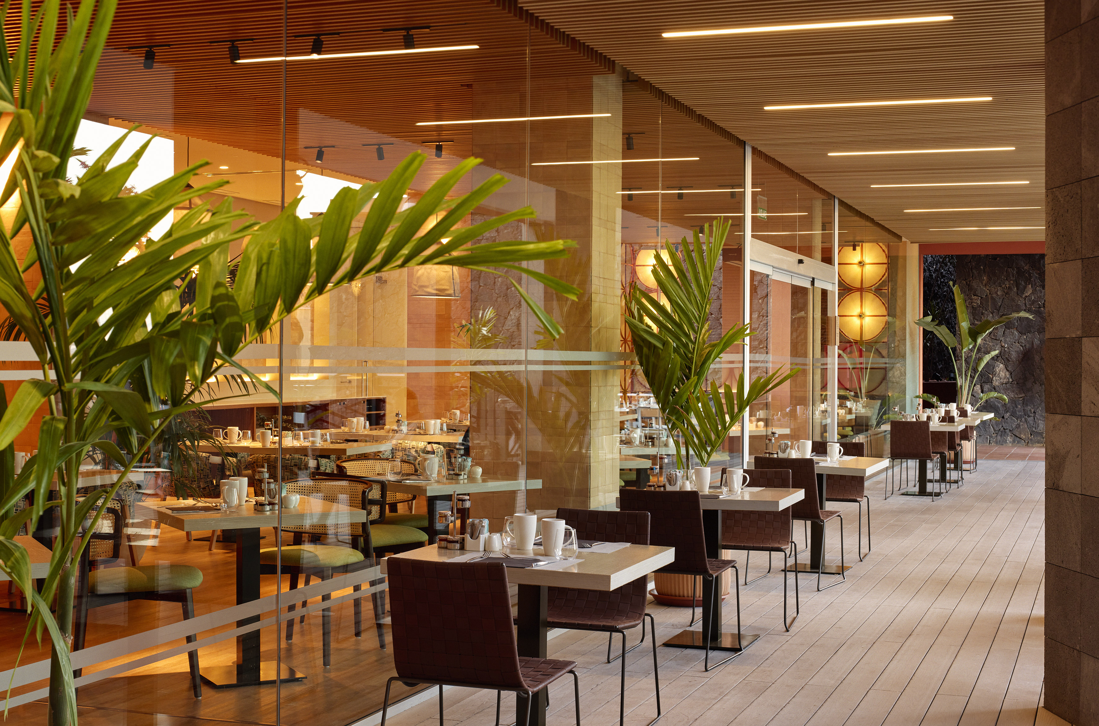 Modern hotel restaurant with glass walls, outdoor dining, and tropical plants.