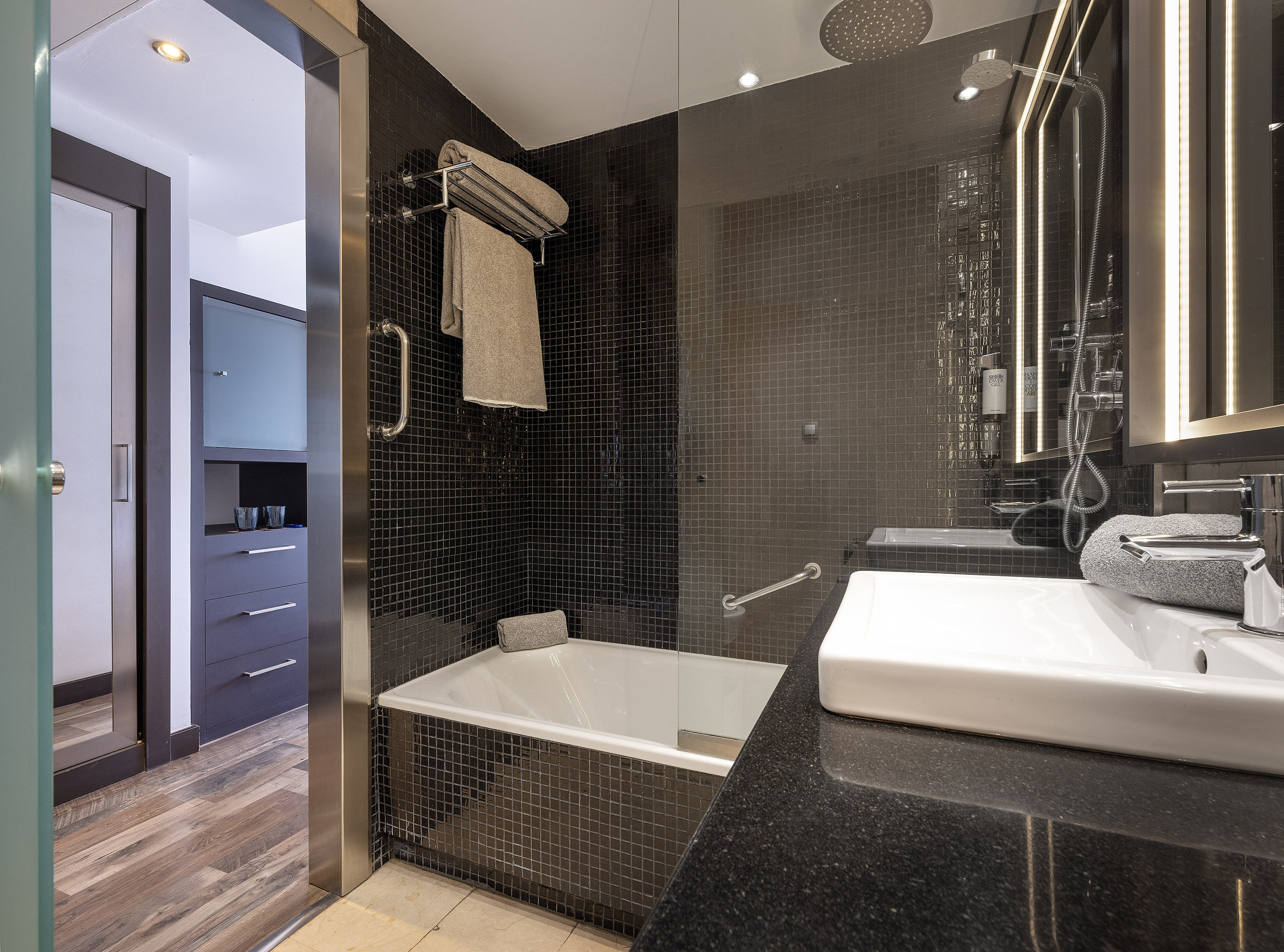 Hotel bathroom with black mosaic tiles, white bathtub, and modern vanity area.