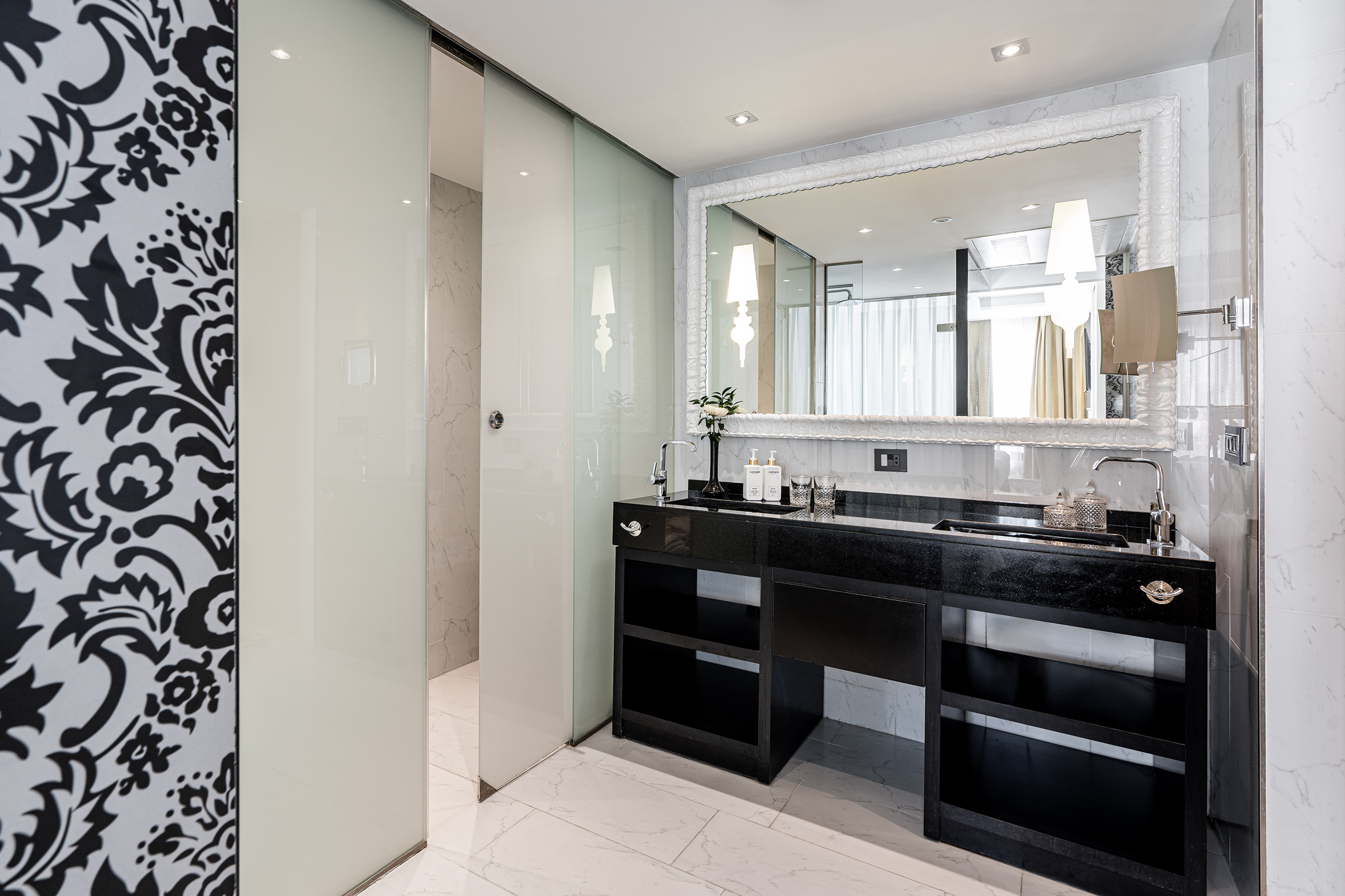 Modern luxury bathroom with ornate mirror, black vanity, glass shower, and damask wall.