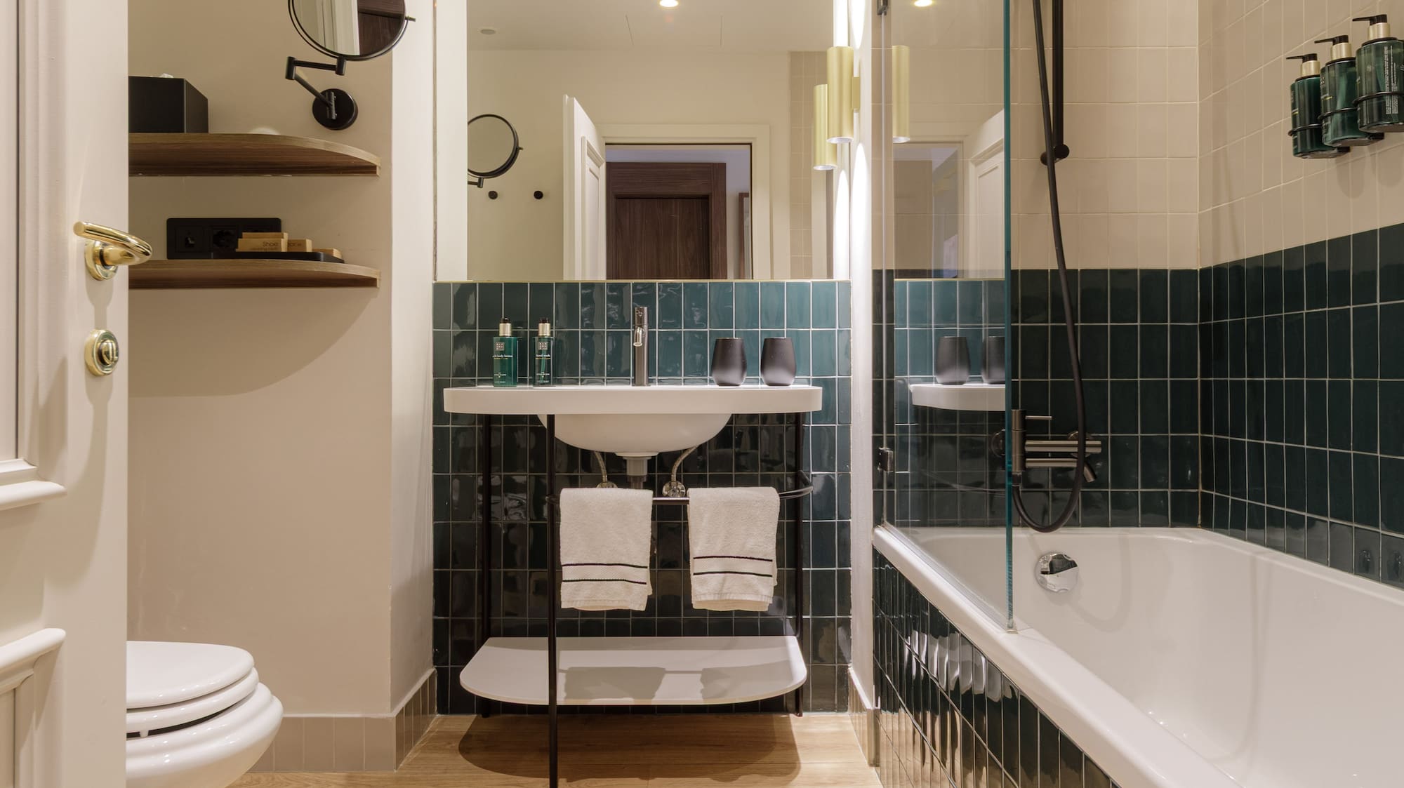 Modern bathroom with wood floor, green tiled walls, white tub, toilet, and sink.