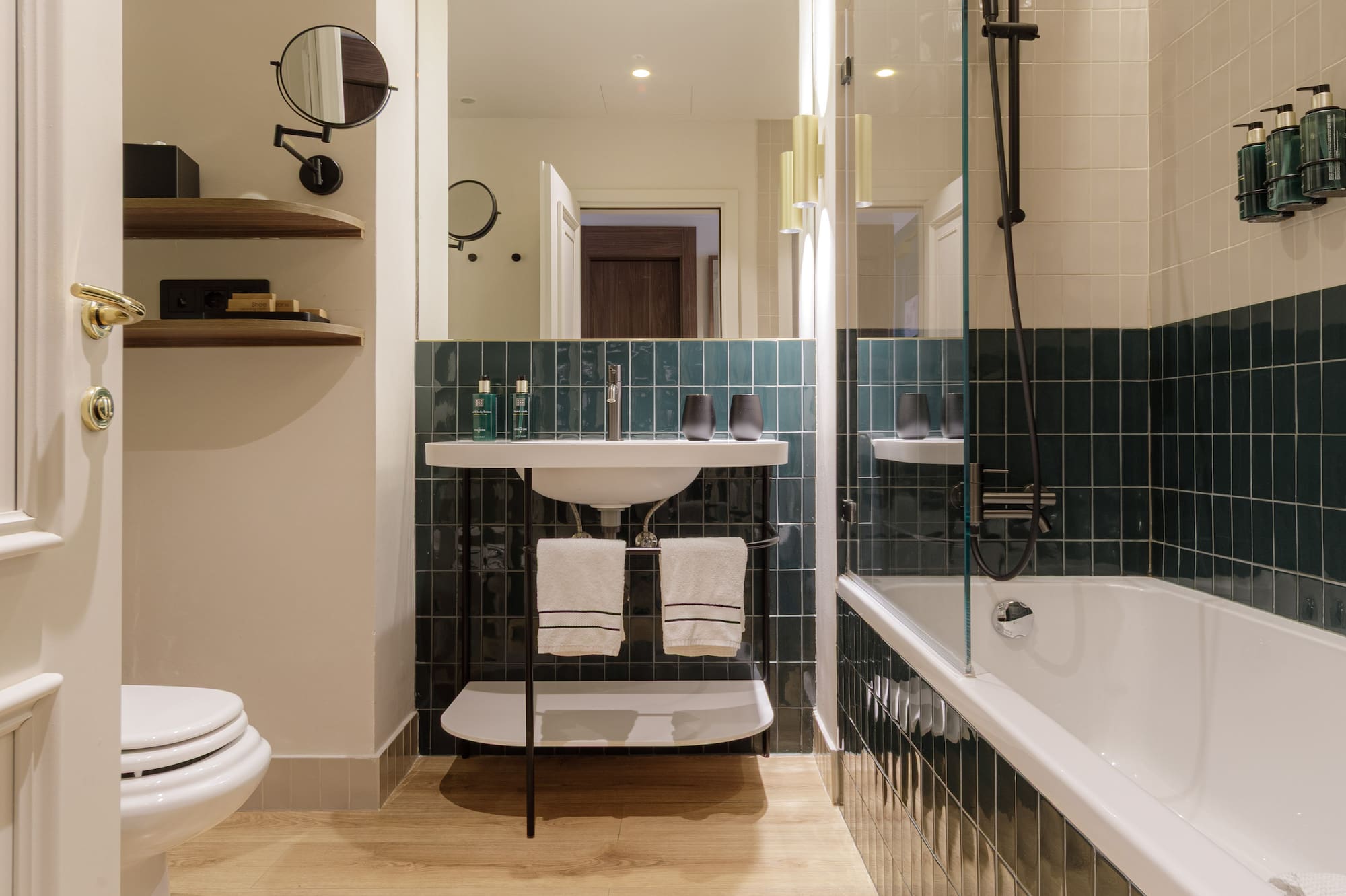 Modern bathroom with wood floor, green tiled walls, white tub, toilet, and sink.