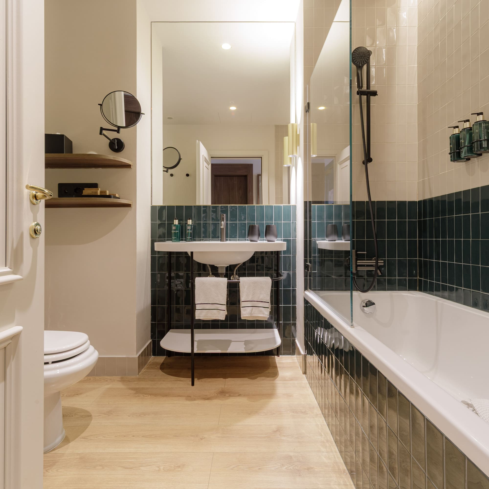 Modern bathroom with wood floor, green tiled walls, white tub, toilet, and sink.