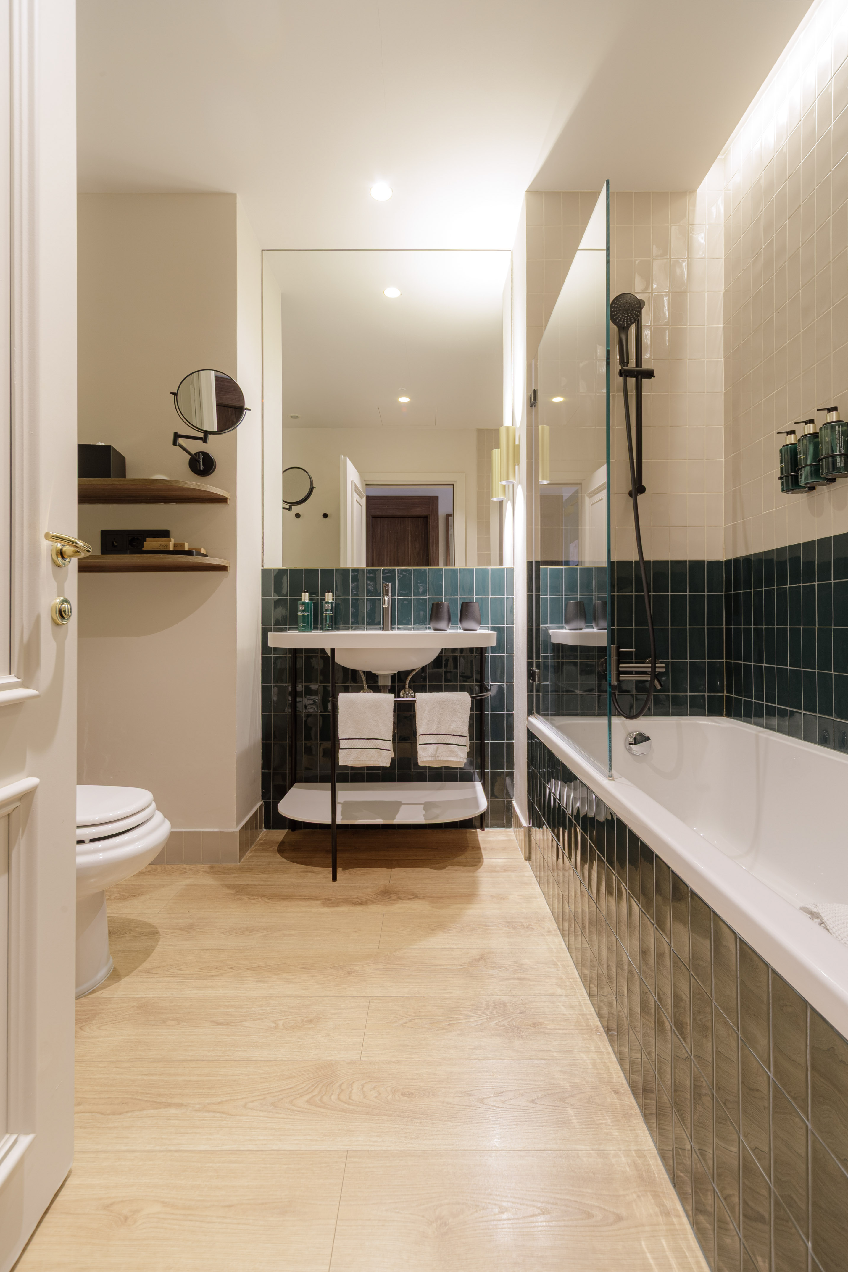 Modern bathroom with wood floor, green tiled walls, white tub, toilet, and sink.