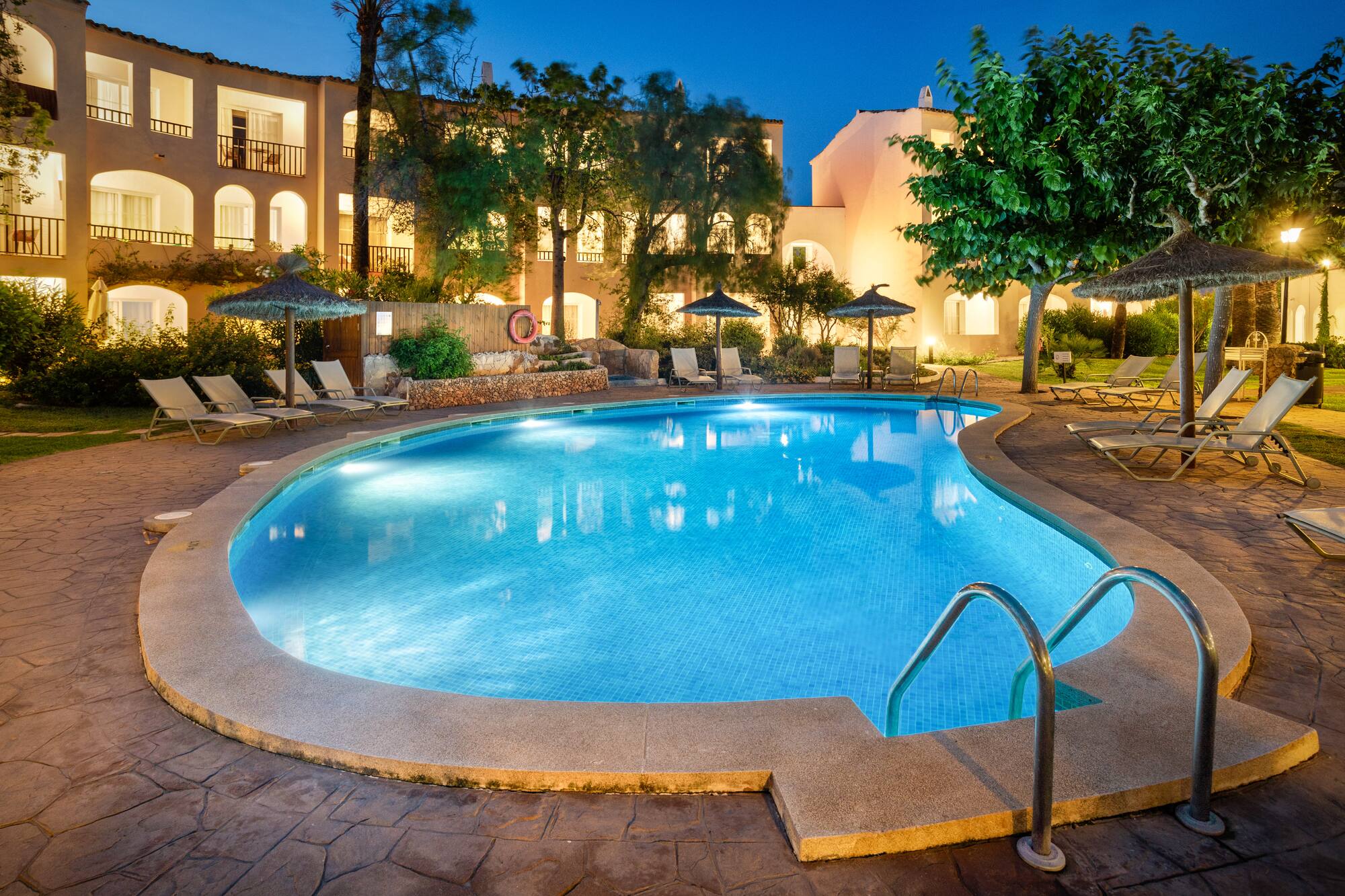 Illuminated resort pool at night with hotel and lounge chairs.