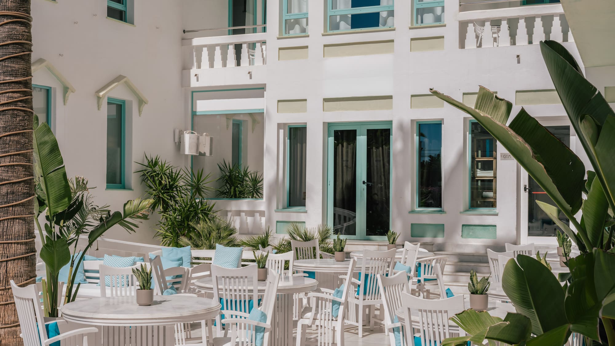 Sunny white resort patio with turquoise accents, dining tables, and tropical plants.