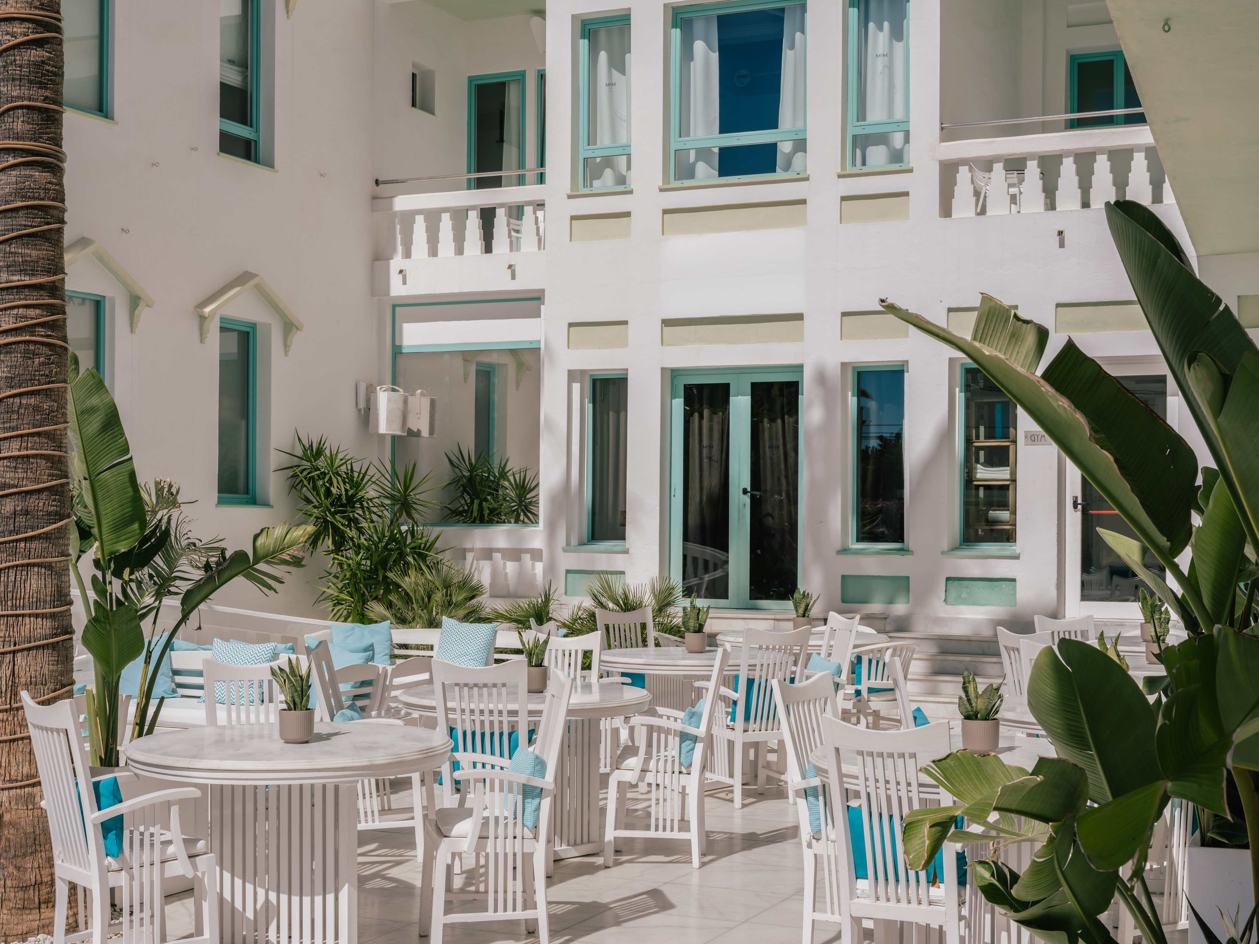 Sunny white resort patio with turquoise accents, dining tables, and tropical plants.