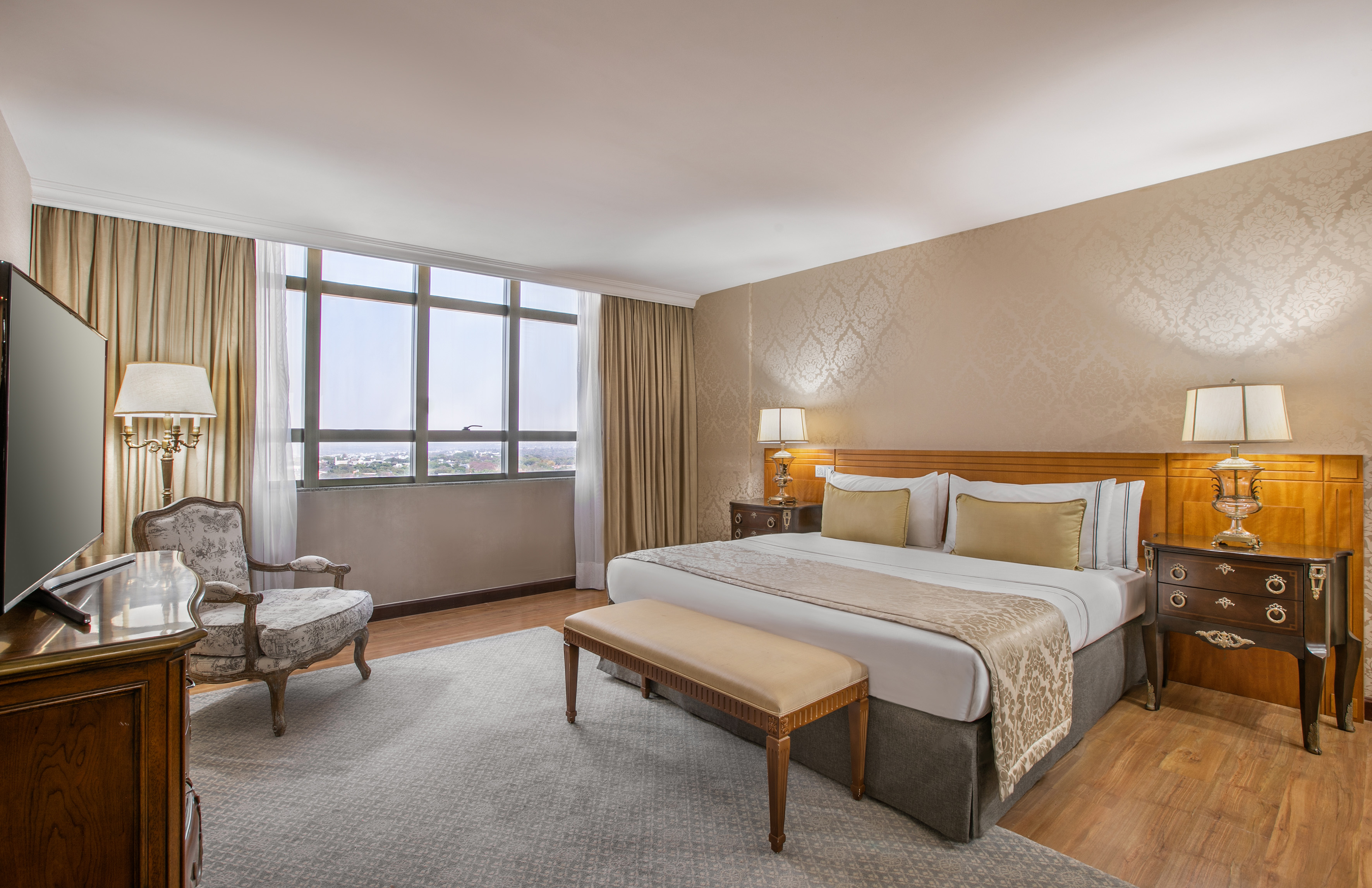 Luxury hotel bedroom with king bed, large window city view, and elegant decor.