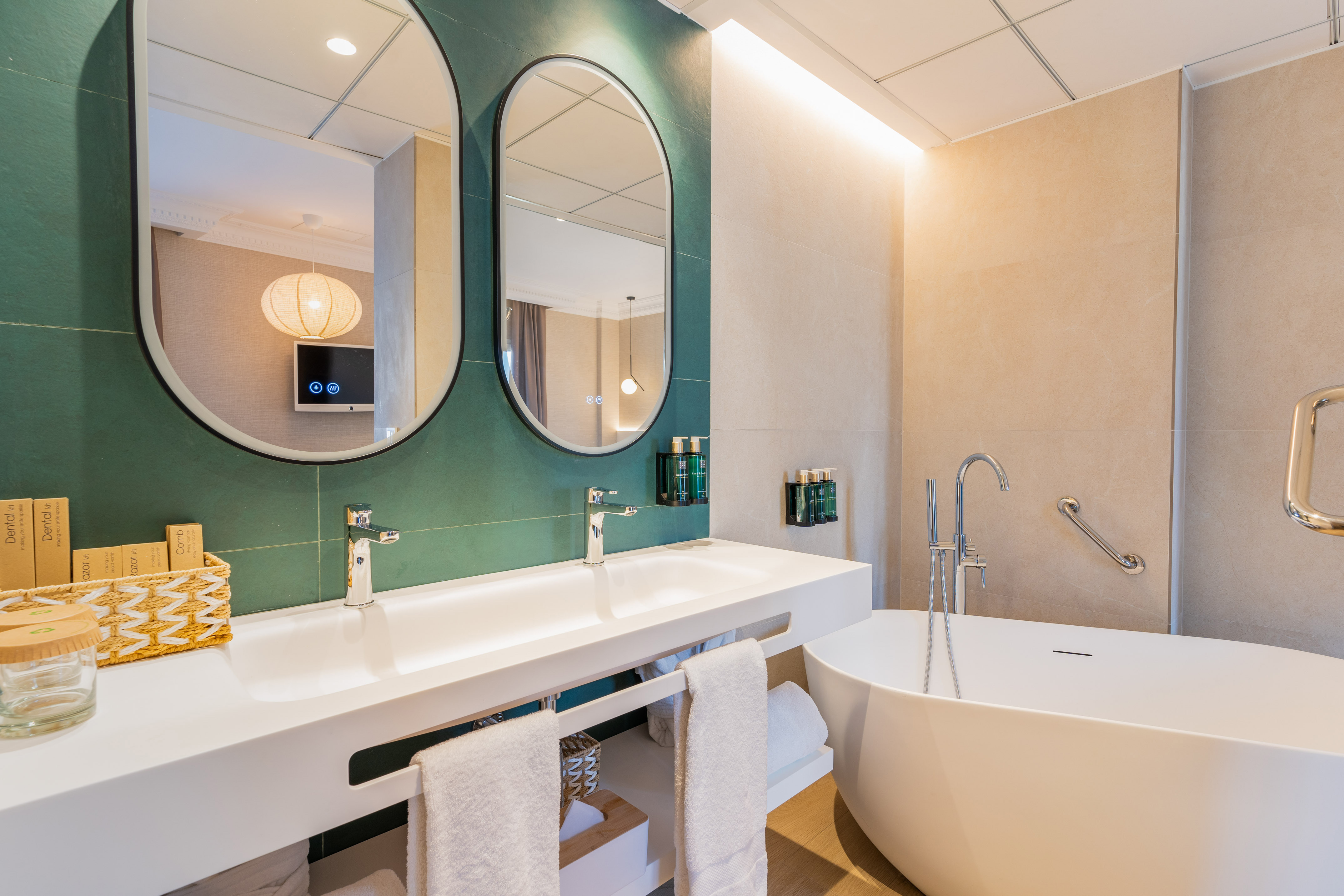 Modern hotel bathroom with double vanity, oval mirrors, green accent wall, and freestanding tub.