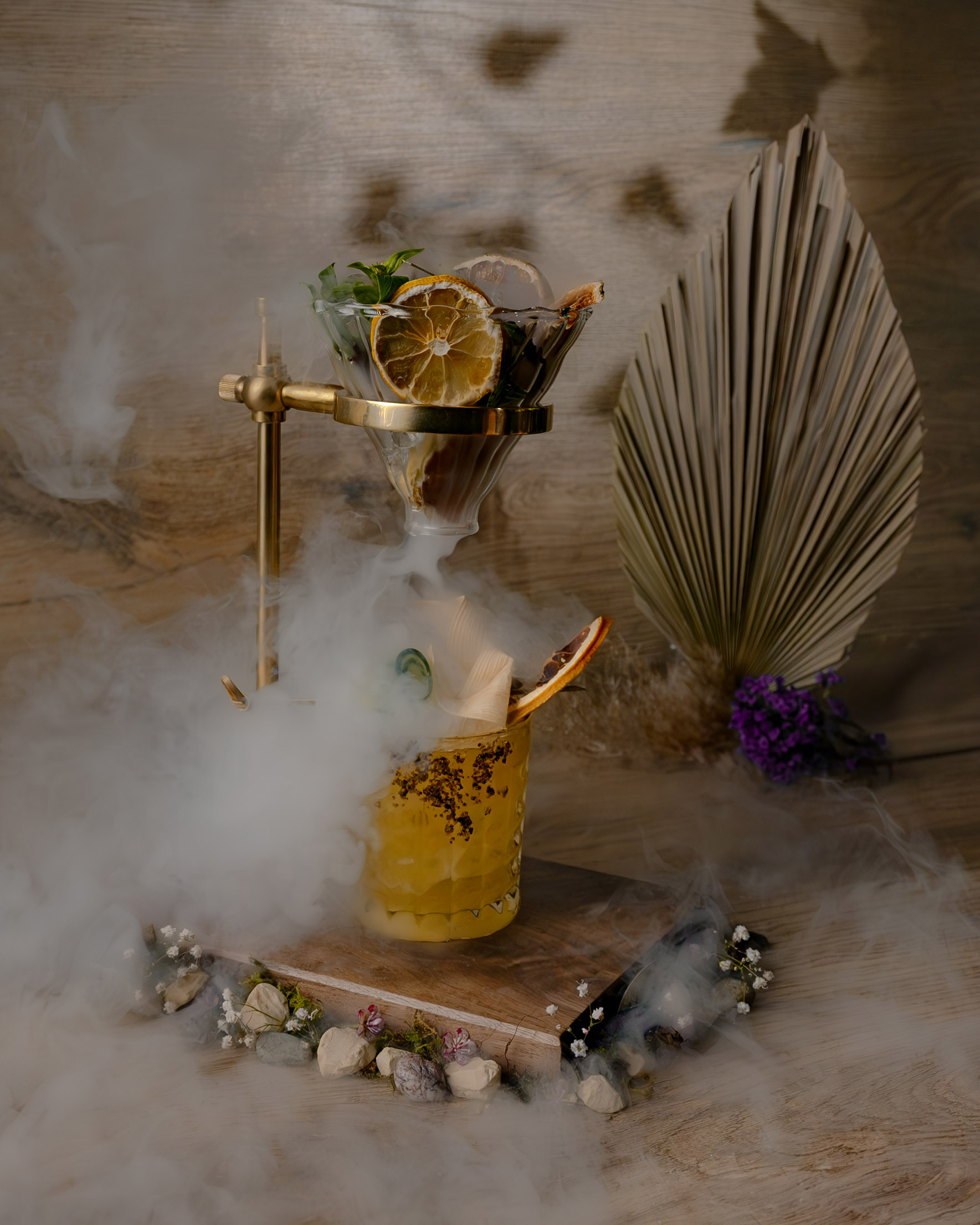 Smoky cocktail with dried orange, mint, golden stand, and mist effect.