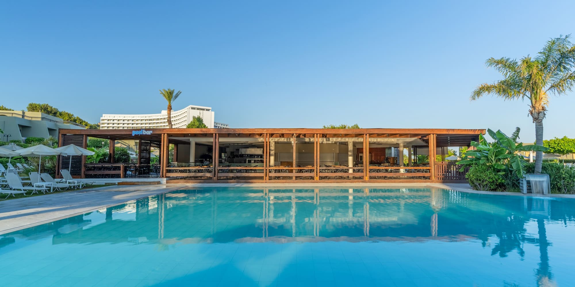Hotel swimming pool and modern pool bar under clear sky with palm trees.