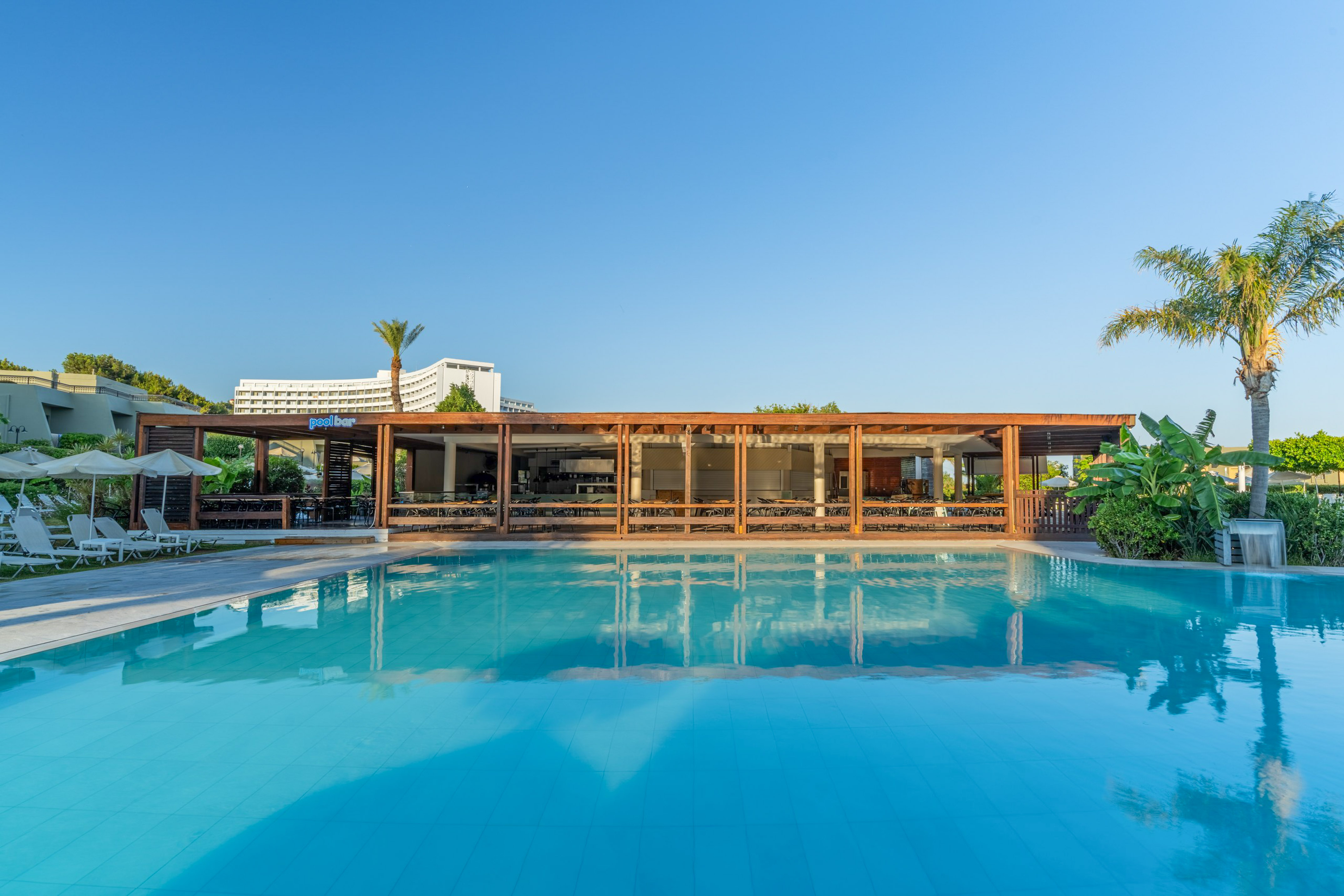 Hotel swimming pool and modern pool bar under clear sky with palm trees.