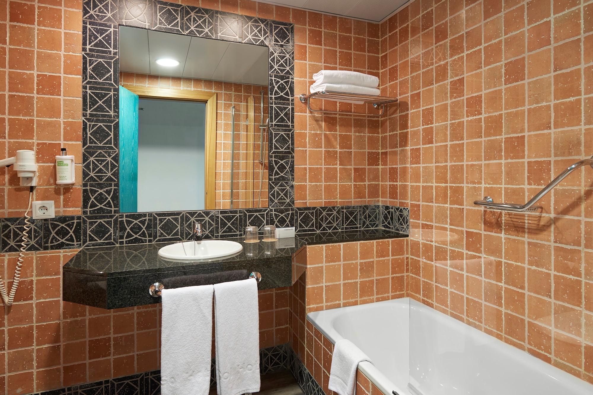 Terracotta tiled hotel bathroom with white bathtub, vanity, and mirror.