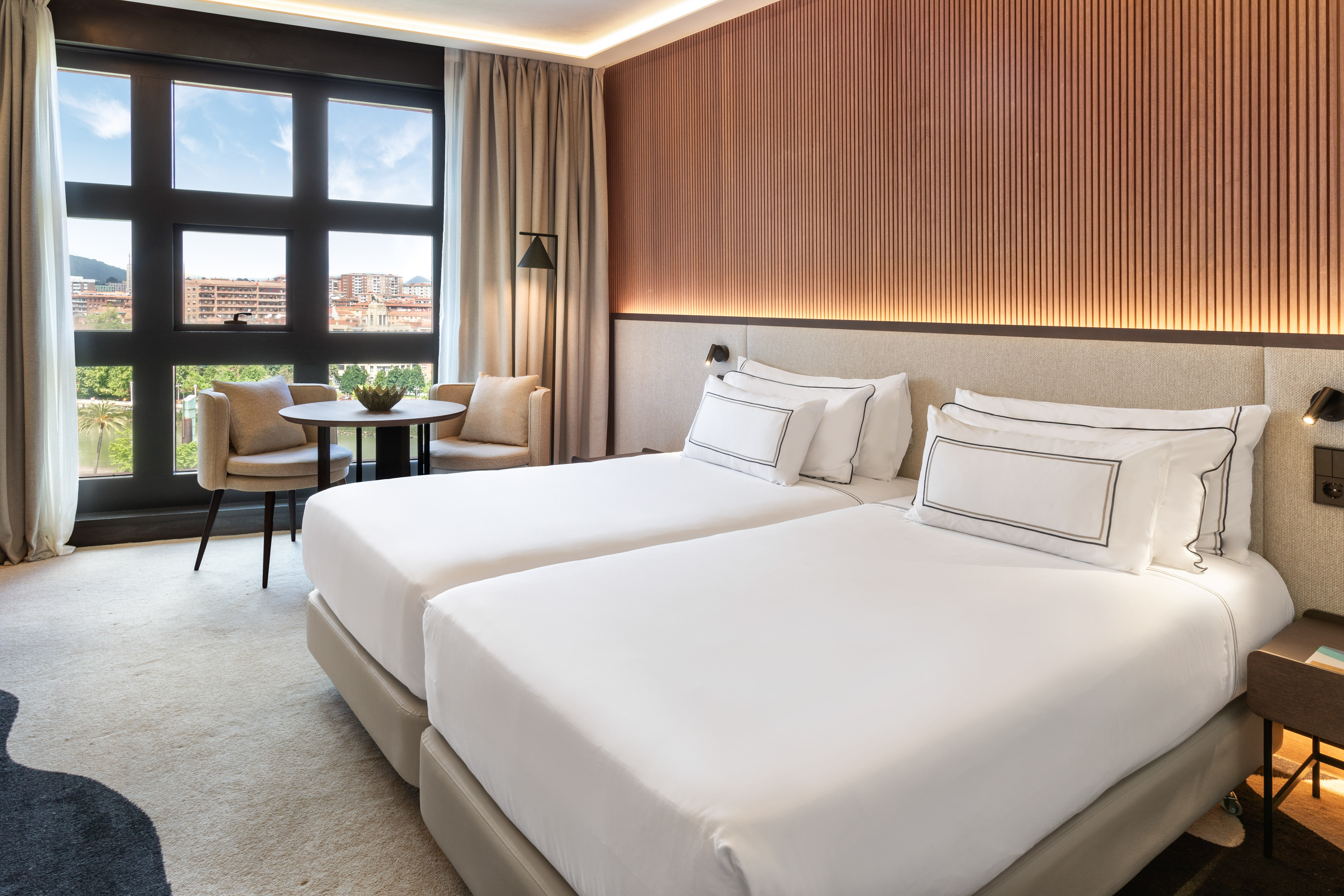 Modern hotel room with twin beds, large window city view, and wood accent wall.