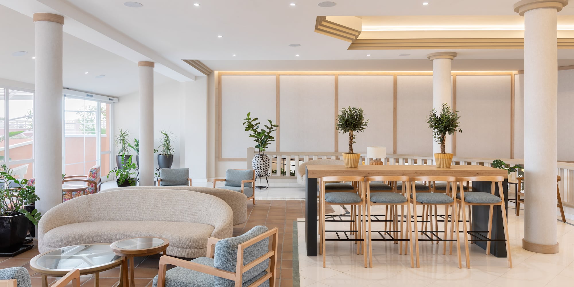 Bright hotel lobby with lounge seating, wooden bar table, and potted plants.