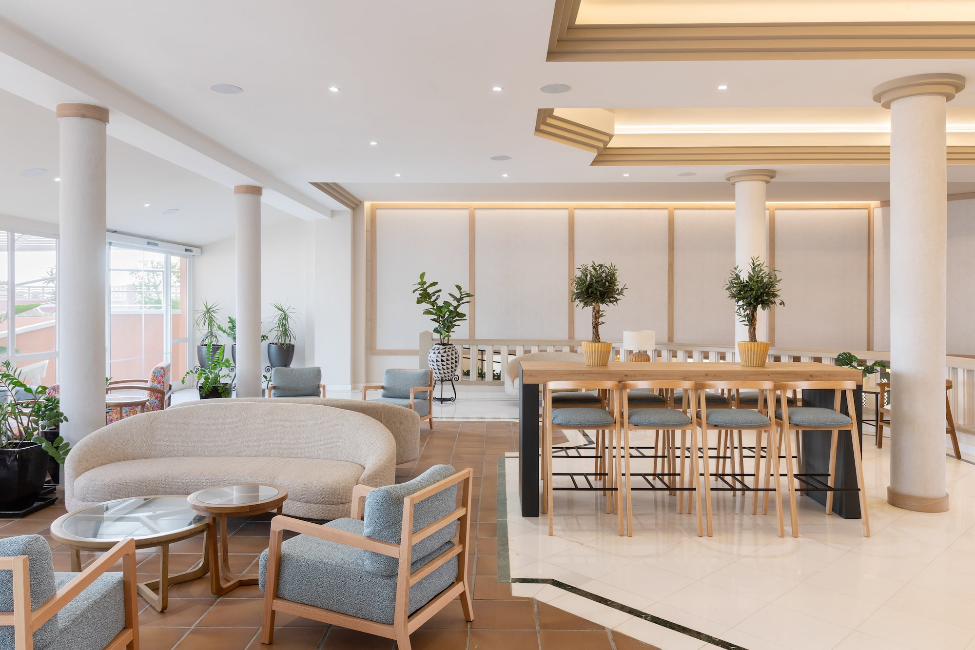 Modern hotel lobby with beige sofa, blue chairs, wooden bar table, and plants.