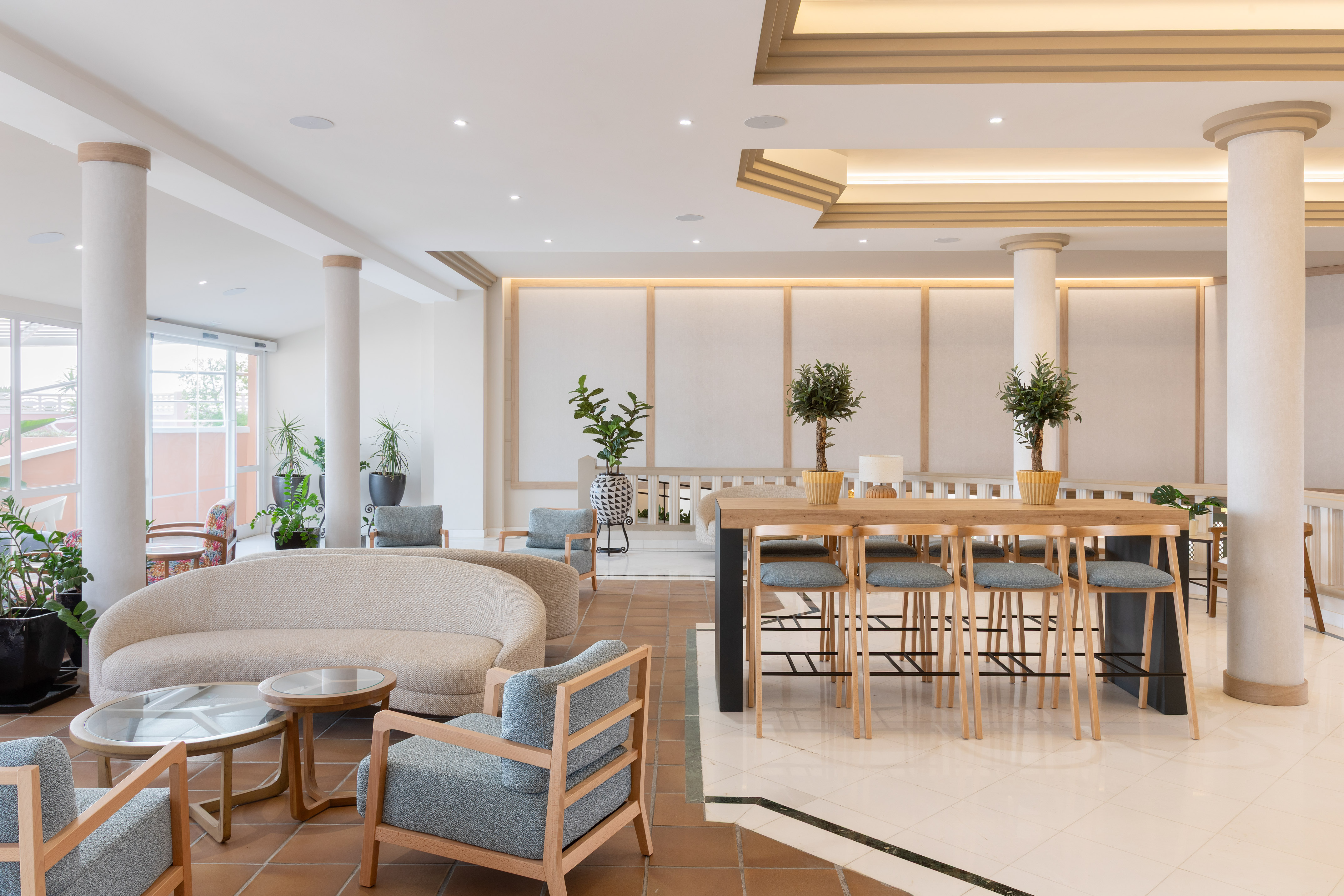 Modern hotel lobby with beige sofa, blue chairs, wooden bar table, and plants.