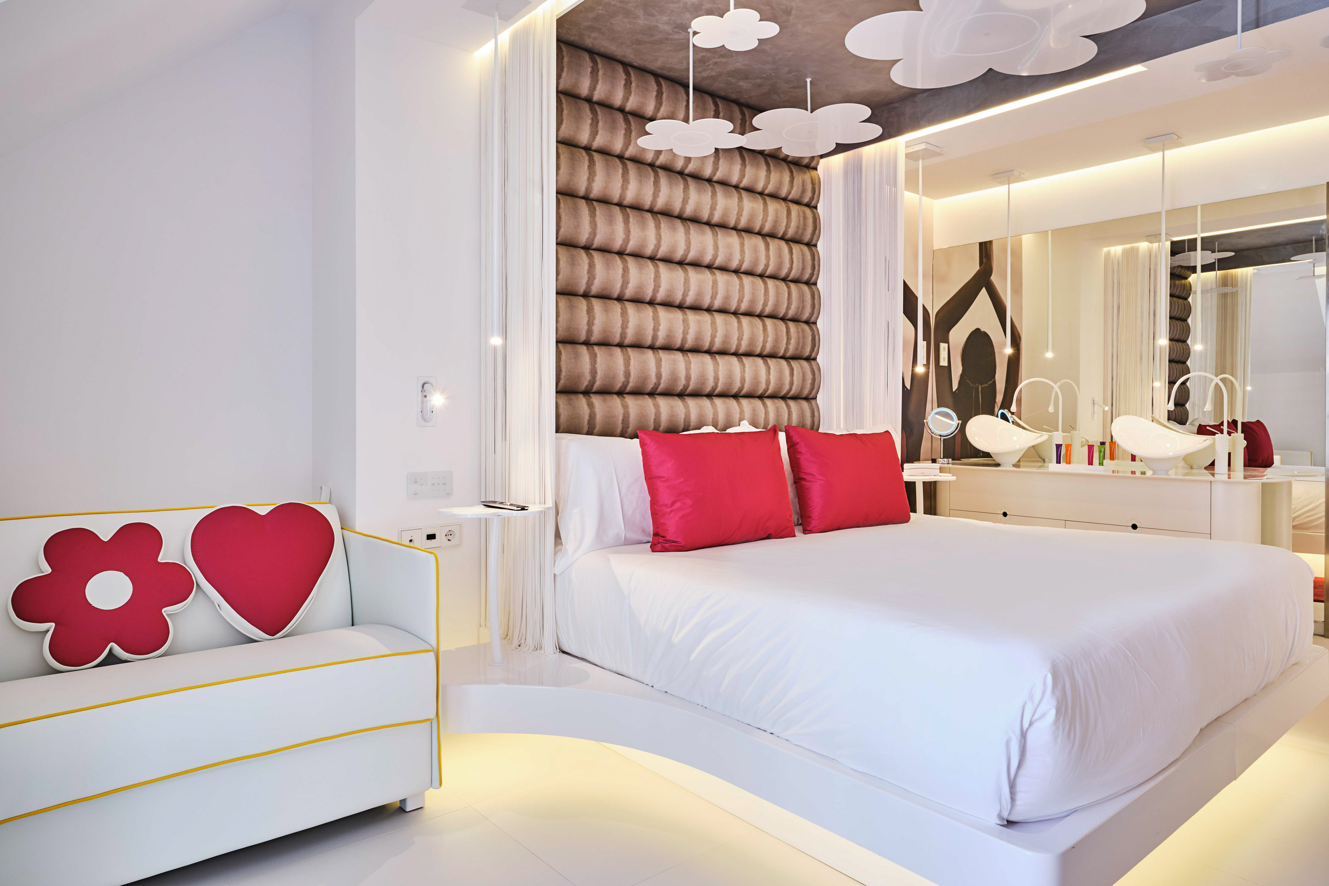 Modern hotel room with white bed, red pillows, cloud decor, and integrated vanity.
